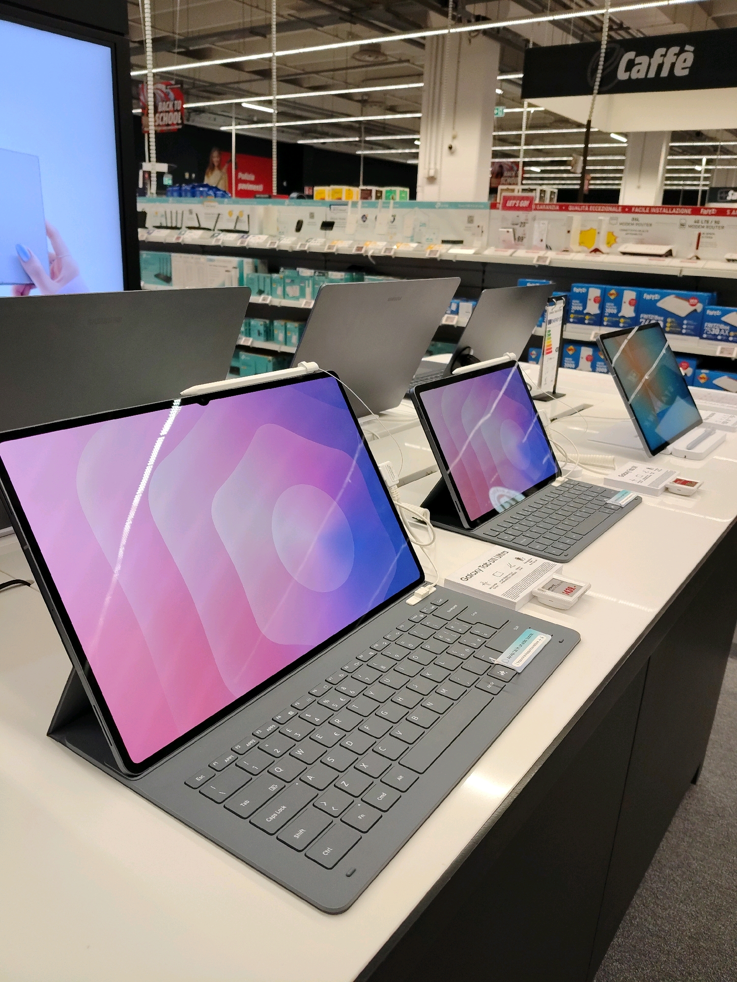 In an electronics store, multiple tablet devices with keyboards are displayed on a white counter. The devices are open and show colorful abstract wallpapers on their screens. One tablet, prominently in the foreground, is a dark gray device with a keyboard. A stylus pen is attached to the top of its screen. Another tablet, to the right, also has a keyboard and a colorful display. In the background, shelves are filled with various electronic products, including routers and other accessories. Signage for "Caffè" is visible above the merchandise. The store lighting illuminates the products on display.
