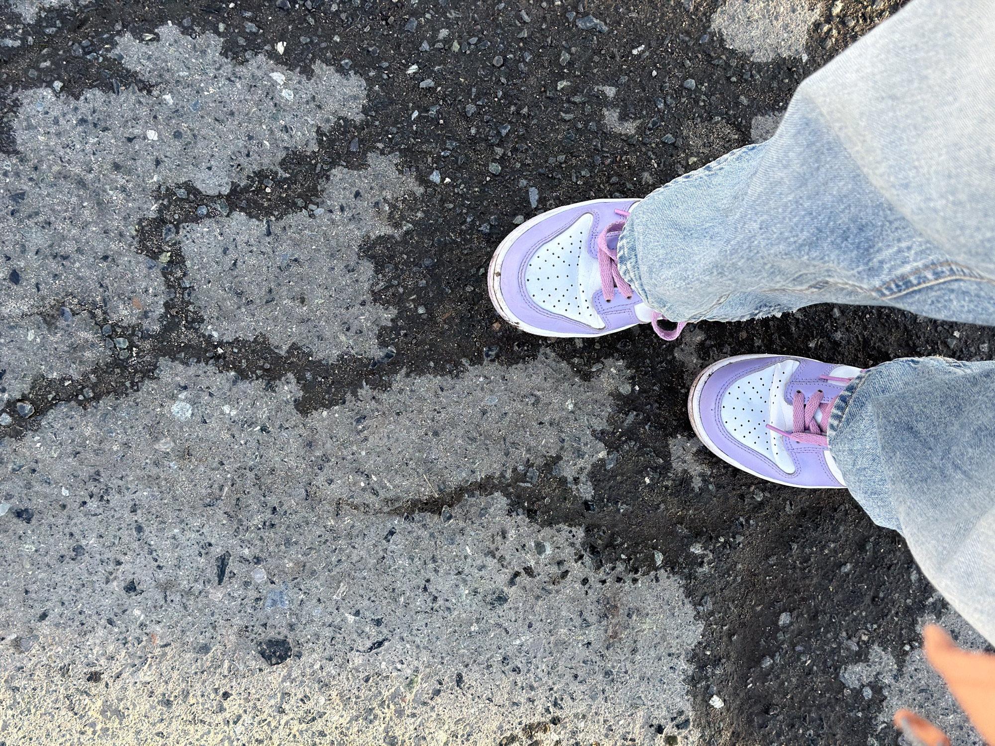 The image presents a top-down view of a person's lower legs and feet, standing on an outdoor ground surface. The individual is wearing light-wash denim jeans, which are slightly bunched around the ankles. On their feet are a pair of sneakers featuring a distinctive purple and white color scheme, accented with pink laces. The sneakers appear to be a mid-top style.
The ground beneath is a textured mix of dark asphalt and lighter grey concrete or pavement. Irregular, darker patches are visible on the asphalt, suggesting areas of moisture or staining, which create a varied pattern across the surface. The lighting indicates it is daytime. The overall setting is an outdoor, possibly urban or suburban environment, located in Berezniki, Russia. No specific activity beyond standing or walking is depicted, and no faces or explicit emotions are visible. There is no discernible text within the image.
