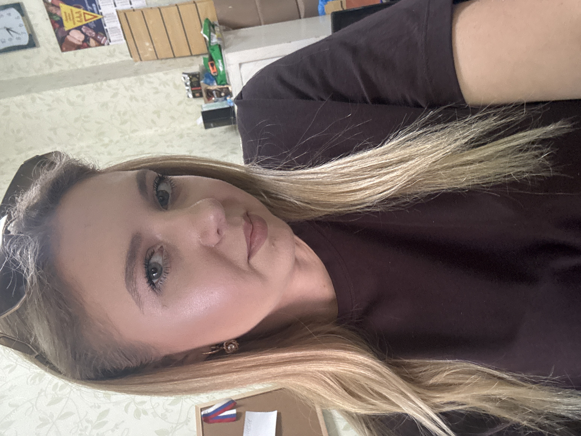 A blonde woman wearing a dark t-shirt takes a selfie in an indoor setting, possibly an office. She has on large sunglasses pushed up onto her head, and she is wearing hoop earrings with a decorative element. Her hair is blonde and falls past her shoulders. In the background, there is a corkboard with a piece of paper and ribbons attached. A clock is visible on the wall, along with a calendar featuring images of food. Shelving units are also present, with various items on them, including boxes and what appear to be rolls of tape. The lighting suggests it may be daytime.
