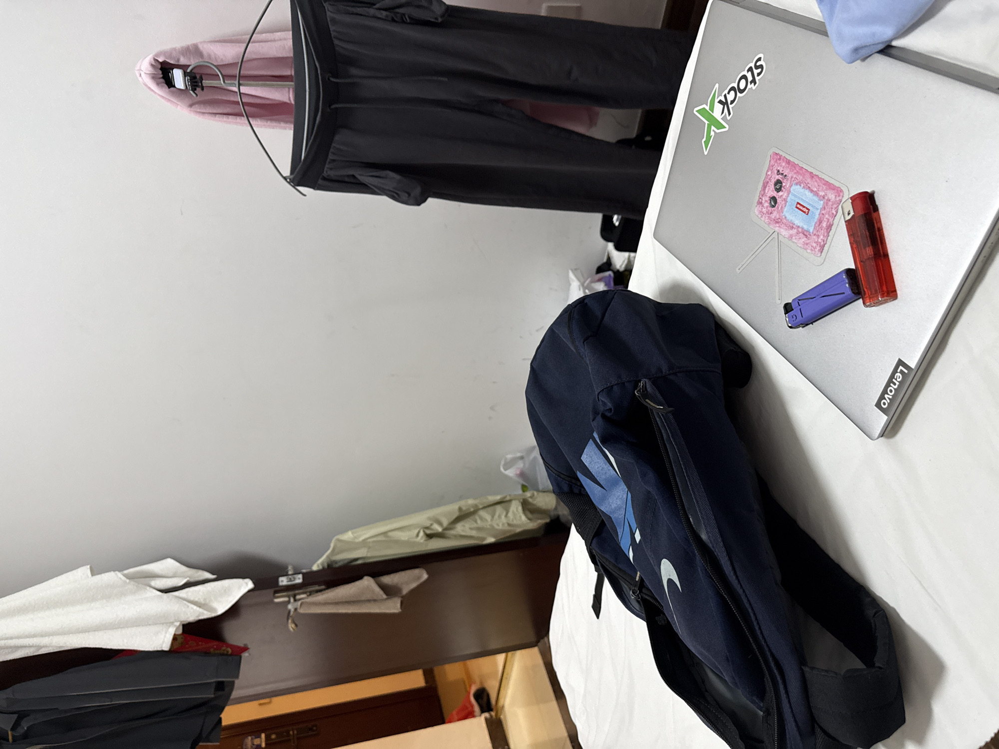 The image depicts a bedroom scene with a bed in the foreground, covered with a white sheet. On the bed, a dark blue backpack is partially open, revealing some contents. To the right of the backpack lies a silver laptop with several stickers on its lid, including a green "StockX" logo and a pink fluffy sticker with a red "Supreme" box logo. Two lighters, one purple and one red, are placed on the laptop. In the background, clothing items are hung on a rack and on hangers against a white wall. A dark wooden door is visible to the left, with more clothing items hanging near it. The overall environment suggests a casual and somewhat cluttered personal space, consistent with a residential setting possibly located in Haizhu District, China, based on the provided context. There are no people visible in the image.