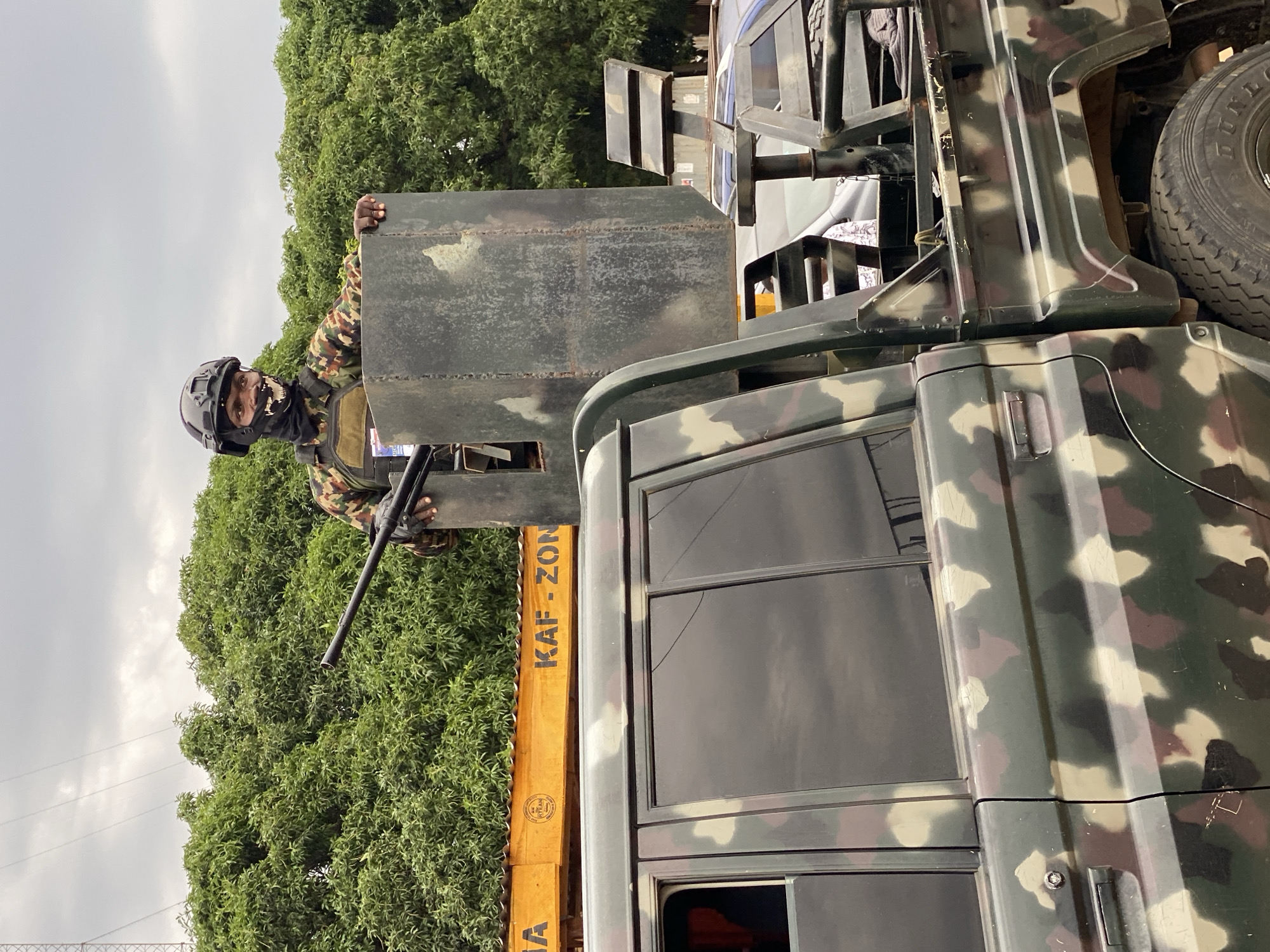 A soldier in camouflage uniform and helmet, possibly a member of KAF-ZON (as indicated on the vehicle), stands atop a camouflage-painted military vehicle. The soldier is positioned behind a makeshift metal shield, holding a firearm. The background shows green trees and an overcast sky. The location is the city of Maigizo, Nigeria.