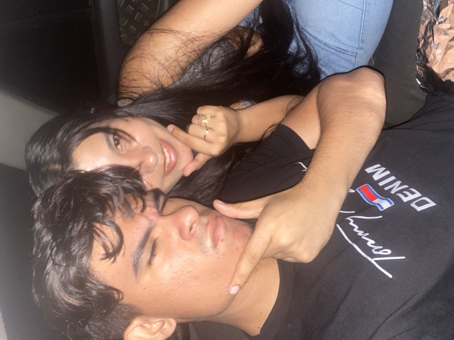A young woman with long dark hair smiles, wearing light blue jeans, while a young man lies with his eyes closed, wearing a black t-shirt with a white "Tommy" logo. The woman is playfully poking the man's cheek with her finger. The photograph is taken at an angle, likely inside a car. The lighting is dim, creating a cozy or intimate atmosphere. The focus is on the faces of the two individuals.