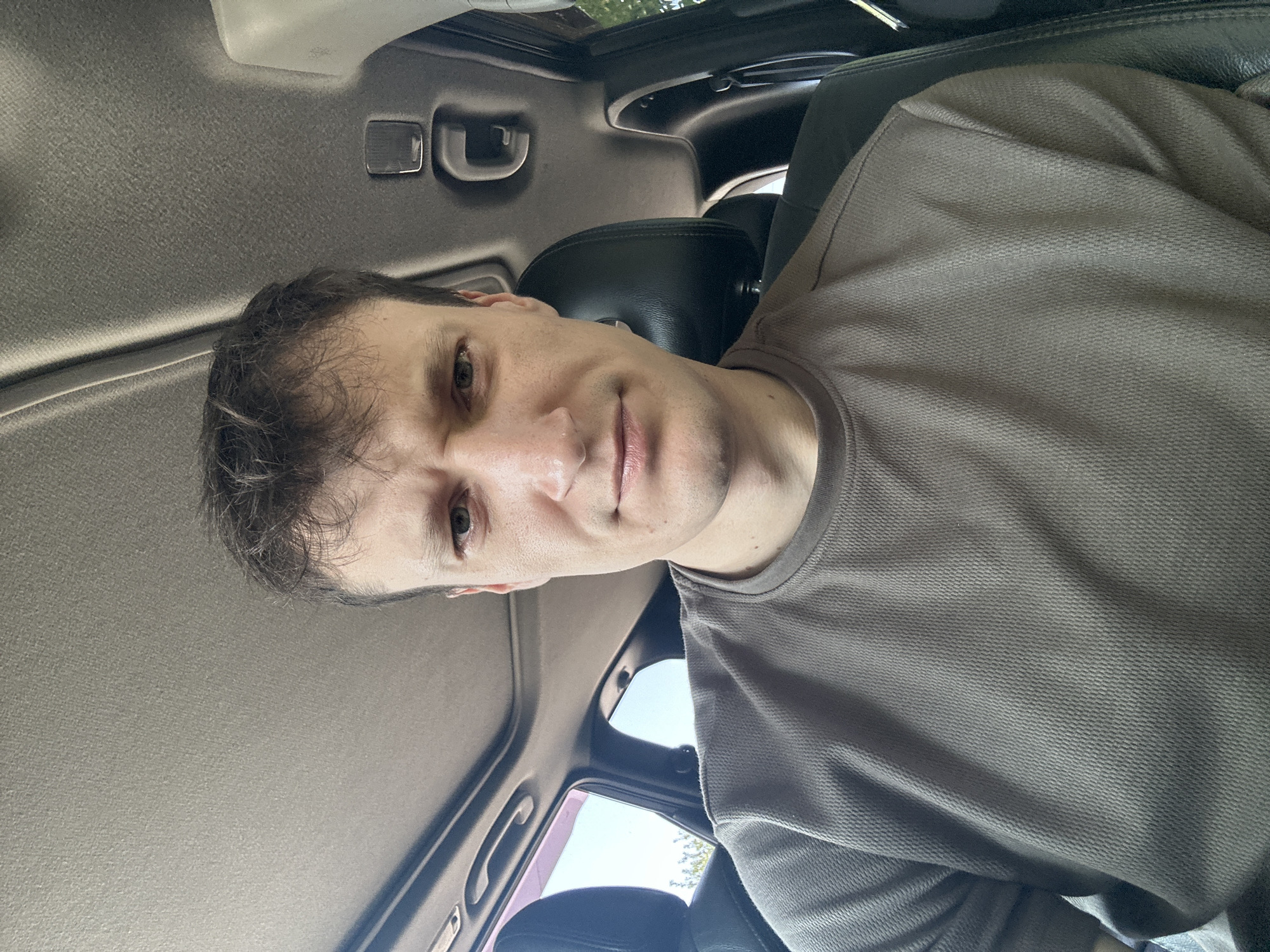 A man with short, messy brown hair and light skin is seated in the driver's seat of a vehicle. He is wearing a brown t-shirt with a textured pattern. His facial expression is one of mild displeasure, with his brow furrowed and lips slightly pursed. The interior of the car is visible, including the grey headliner, a sunroof, and a black headrest. The outdoor environment is visible through the car windows, suggesting a daytime setting. The image was taken in Makiv, Ukraine.