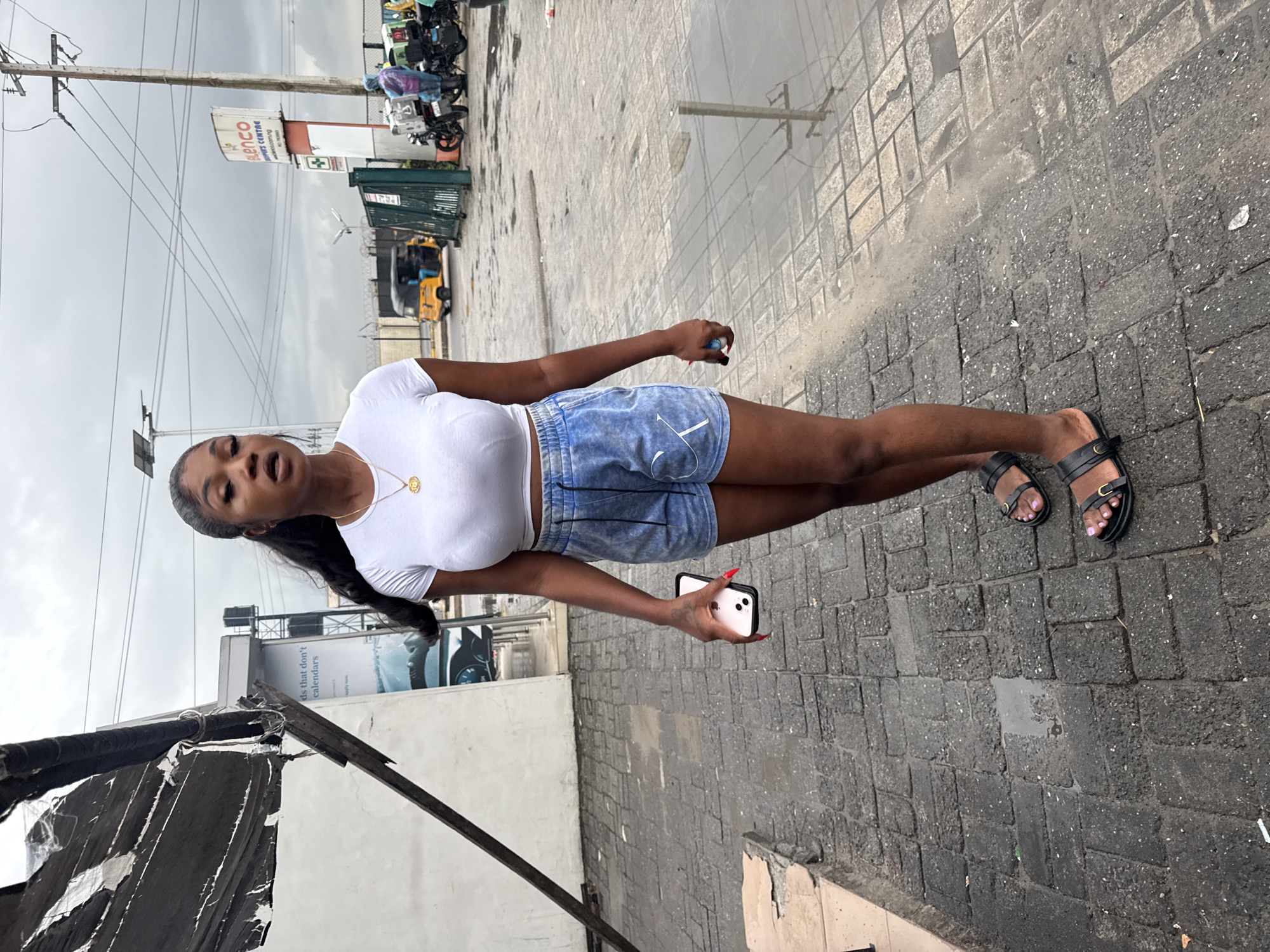 A full-body, outdoor, eye-level shot captures a young Black woman standing on a gray, patterned brick sidewalk in what appears to be an urban environment. She is wearing a white, short-sleeved, form-fitting crop top, blue denim shorts with white drawstrings and a white logo, and black sandals. Her dark hair is pulled back into a high ponytail, and she is wearing a gold necklace with a circular pendant. In her right hand, she holds a white smartphone. The background features a white building with a black tarp covering part of its roof, a billboard with text including "BLENCO SHOPPERS CENTRE" and a website address, electrical poles and wires, a yellow auto-rickshaw, motorcycles, and a few individuals dressed in casual attire. The ground is wet, indicating it may have recently rained, with puddles reflecting the sky and surrounding objects. The overall atmosphere suggests a cloudy day in a commercial or public area.