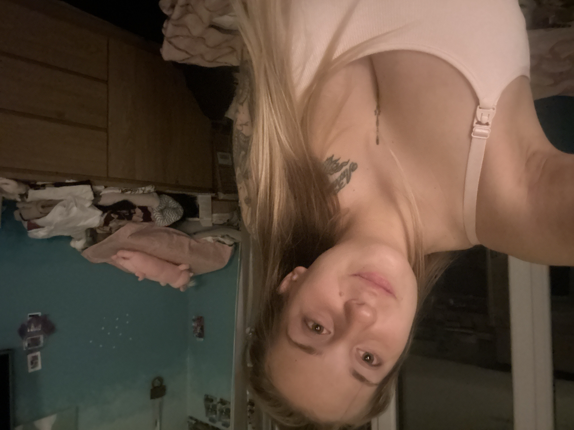 A young woman with blonde hair, wearing a light pink tank top, is taking a selfie in what appears to be a bedroom. She has a tattoo on her chest and arm. The room is decorated with turquoise walls and has a wooden dresser in the background, which is cluttered with various items. There is also a pink stuffed animal visible on top of the dresser. The lighting suggests it is evening or a dimly lit room. There is no indication of the location being in Plovdiv, Bulgaria.