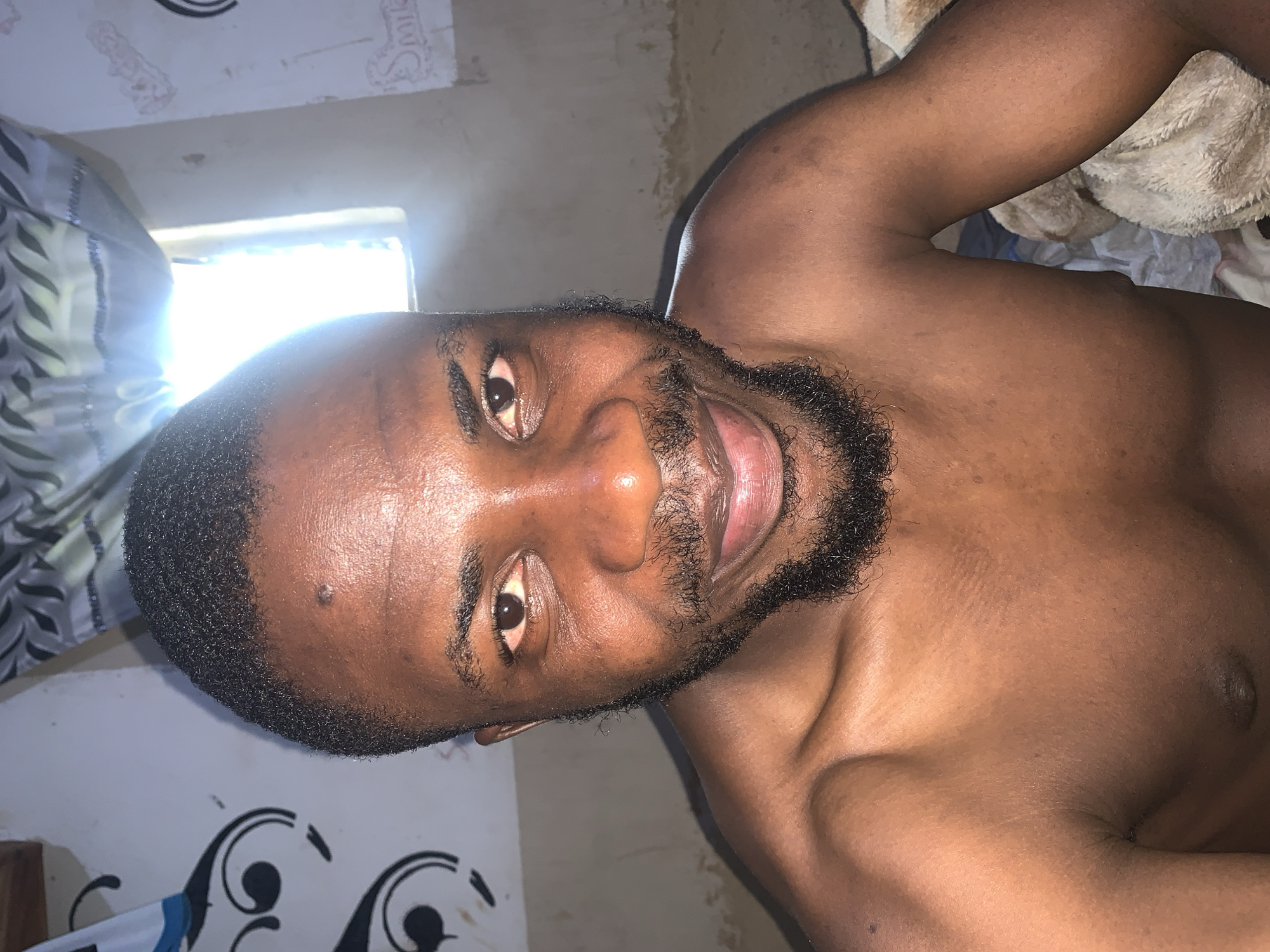 A close-up, eye-level, head and shoulders shot shows a Black man with short, dark, wavy hair and a beard. He is looking directly at the camera with a slight smile. He is shirtless, and his chest and shoulders are visible. The background consists of a light-colored wall with some dark decorative swirls on the left and what appears to be a window with bright light coming through on the right. The overall lighting is warm and somewhat dim, with a glare from the window. It is difficult to determine the exact location within Rafin Par, Nigeria, from the image alone, but the setting appears to be domestic, possibly a bedroom or living area.