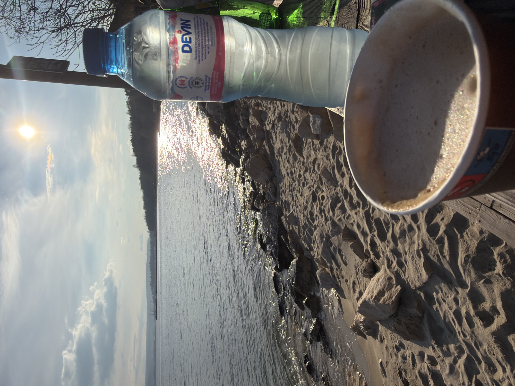 The image captures a serene scene at a beach, likely near Varna, Bulgaria, as indicated by the "DEVIN" bottled water, a popular brand in the country. The foreground features a cup of coffee placed on a sandy surface near the edge of a body of water, presumably the Black Sea. Adjacent to the coffee cup is a plastic bottle of water, and further in the background, there are glimpses of vegetation. The sun is visible in the sky, its reflection shimmering on the water surface. The overall environment suggests a calm morning or afternoon at a seaside location.
