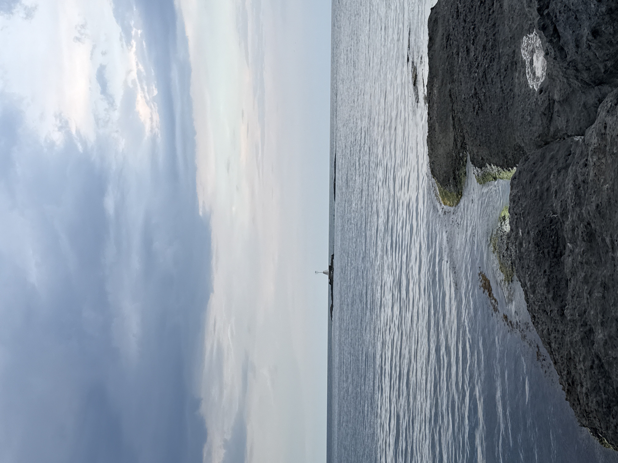 This is a photograph of the Black Sea coast in Tsarevo, Bulgaria. In the distance, a small rocky outcrop with a white structure, possibly a navigational marker or a small lighthouse, is visible on the horizon. The sea is calm with gentle ripples, and the sky is overcast with a mix of dark and lighter clouds, suggesting an impending change in weather or the aftermath of a storm. The foreground is dominated by dark, rough rocks along the coastline, with some patches of green algae visible where the water meets the land. There are no people or animals in the image. The overall mood is serene and somewhat melancholic due to the muted colors and cloudy sky.