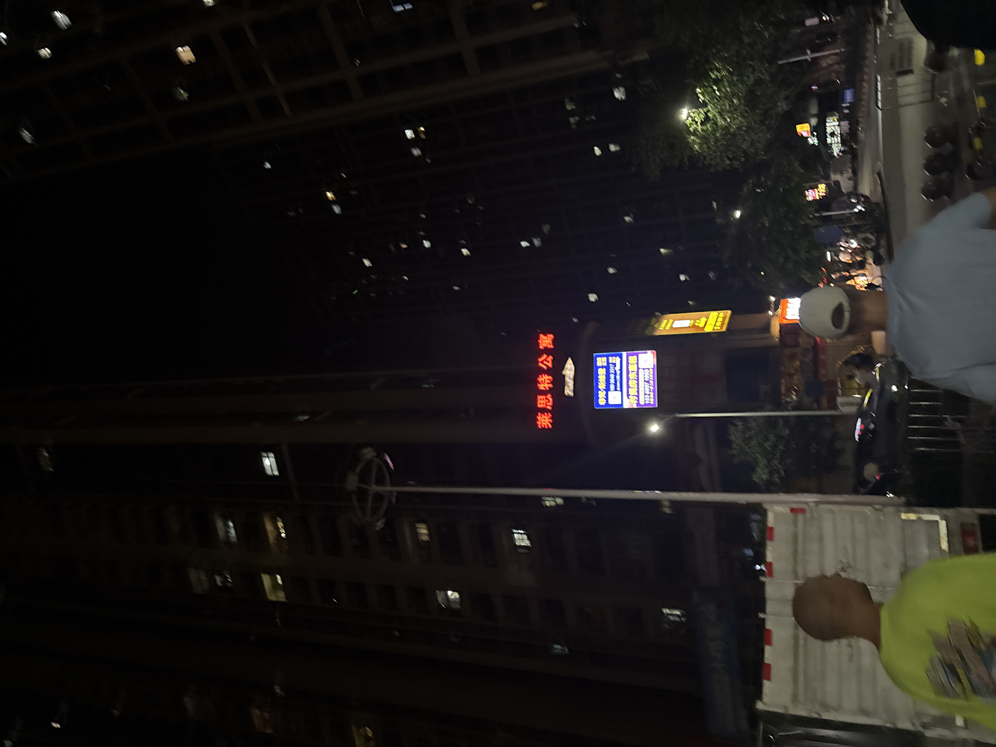 The image depicts a nighttime street scene in Haizhu District, China. The view is dominated by tall buildings with illuminated windows, suggesting an urban environment. In the foreground, the back of a white truck is visible. A person wearing a bright yellow-green shirt is standing next to the truck, facing away from the camera. The overall lighting is dim, typical of nighttime, with streetlights and building lights providing illumination.
In the midground, there is a street with several vehicles, including motorcycles, and pedestrians. A blue and white sign with Chinese text is prominently displayed, along with another illuminated sign with red Chinese characters that reads "惠思特公寓". There are trees lining the street, and more buildings in the background. The perspective is slightly tilted, giving a sense of depth. The scene appears to be bustling with activity, as indicated by the presence of vehicles and people.