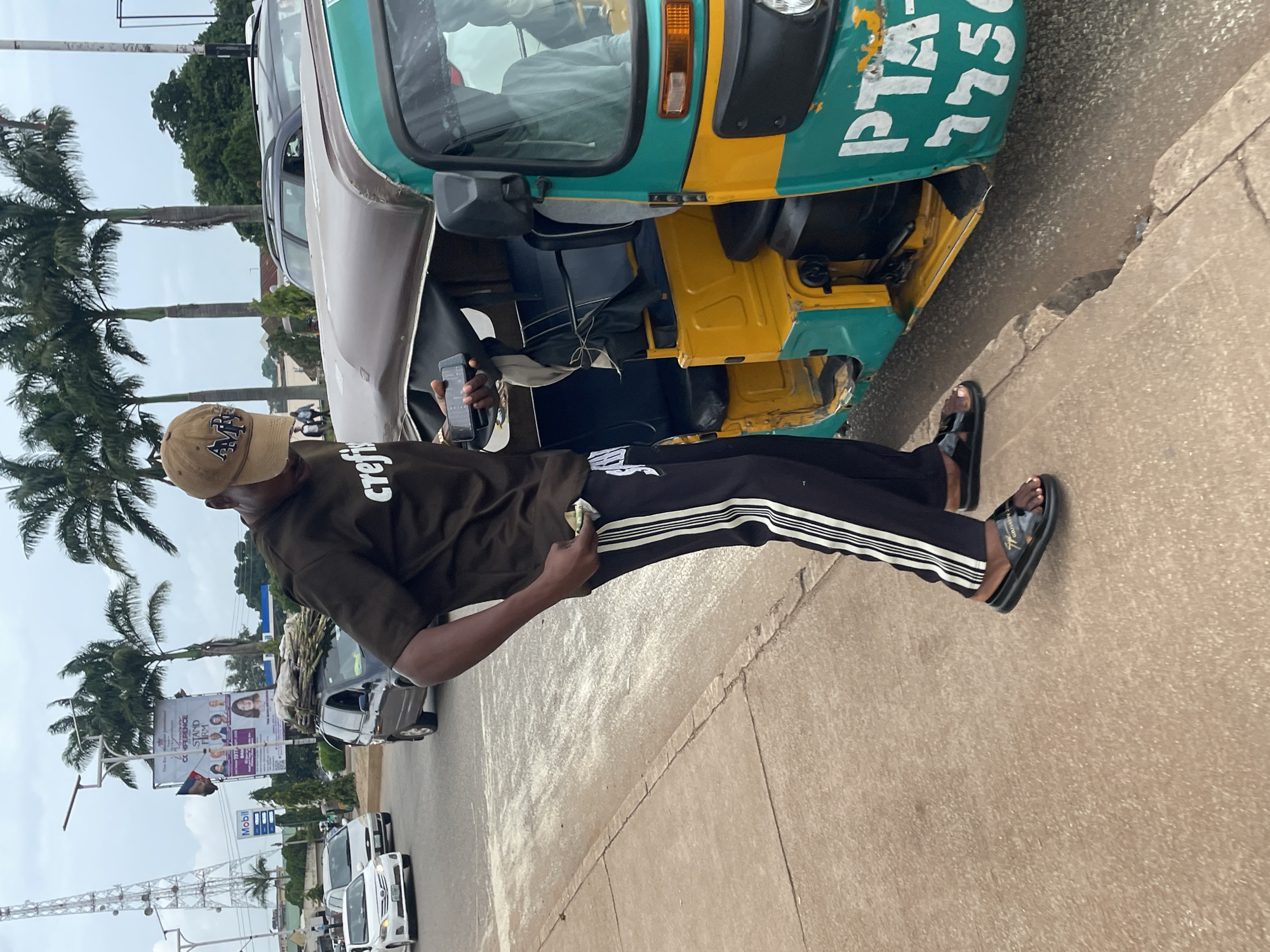 A person is standing on a cracked concrete sidewalk next to a turquoise and yellow auto rickshaw on a street in Jos, Nigeria. The person is wearing a brown t-shirt with white lettering, black pants with white stripes on the sides, a brown baseball cap, and black slide sandals. They are holding a mobile phone in their right hand and have some bills sticking out of their left hand. The auto rickshaw has white lettering on its side that reads "PTA 7750". In the background, there are palm trees, other vehicles, a cell tower, and a billboard advertising a conference. The sky is cloudy.