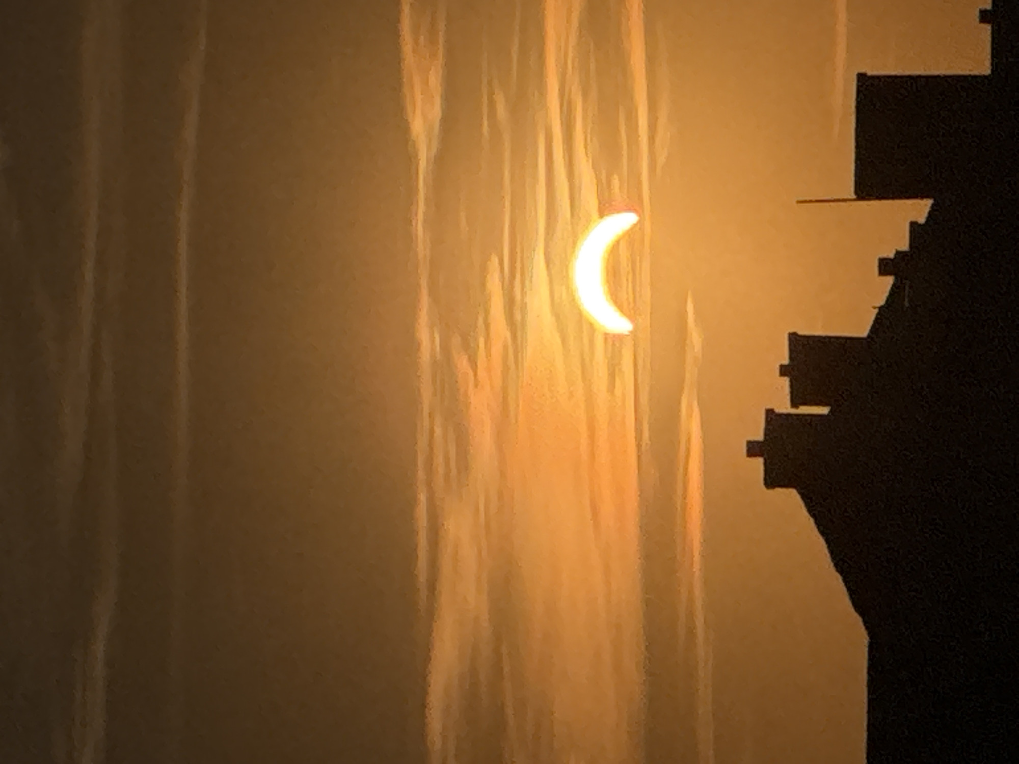 The image captures a partial solar eclipse over Wettstetten, Germany, during what appears to be either sunrise or sunset. The sky is a warm gradient of orange and yellow, with wispy clouds contributing to the atmospheric effect. A distinct crescent shape of the sun is visible, illuminated by its own light. Silhouetted against the glowing sky are the rooftops and chimneys of several buildings, indicating an urban or suburban setting. The foreground is dominated by the dark outlines of these structures, with no people or discernible actions visible. The overall scene is serene and highlights a celestial event against a backdrop of human architecture.