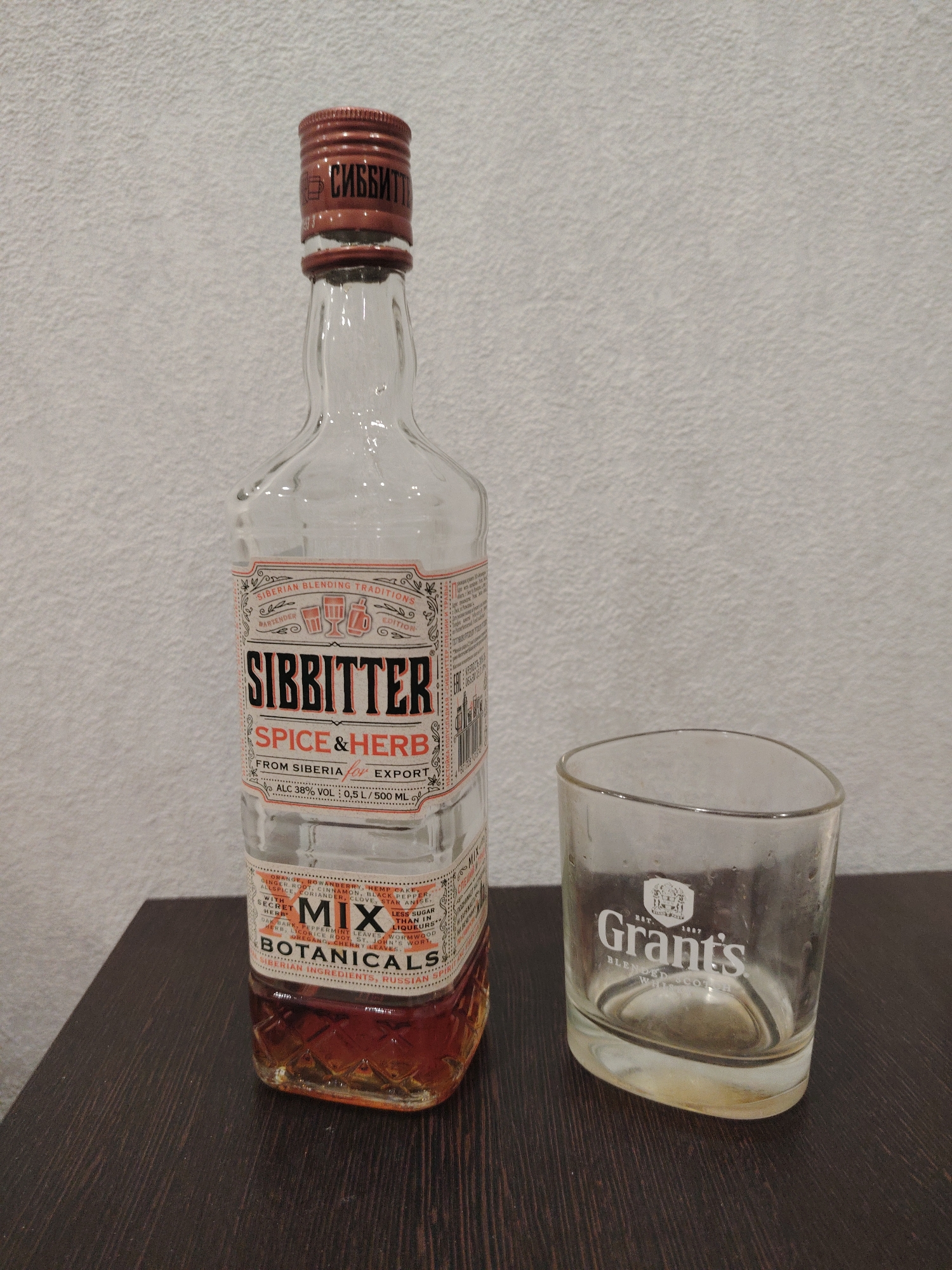 The image shows a nearly empty bottle of "Sibbitter" spice and herb liqueur next to a "Grant's" blended scotch whisky glass. The bottle has a reddish-brown cap with Cyrillic text, and the label indicates it is a "Siberian blending tradition" from Siberia, Russia, with 38% alcohol content. The label lists various botanicals including orange, rowanberry, ginger root, cinnamon, black pepper, allspice, coriander, clove, star anise, oak bark, peppermint leaves, licorice root, St. John's wort, wormwood, oregano, and cherry leaves. The glass appears to be slightly dirty, with traces of liquid at the bottom. They are both placed on a dark wooden surface against a plain, light-colored wall. The image does not depict the city of Sarapul or any people.