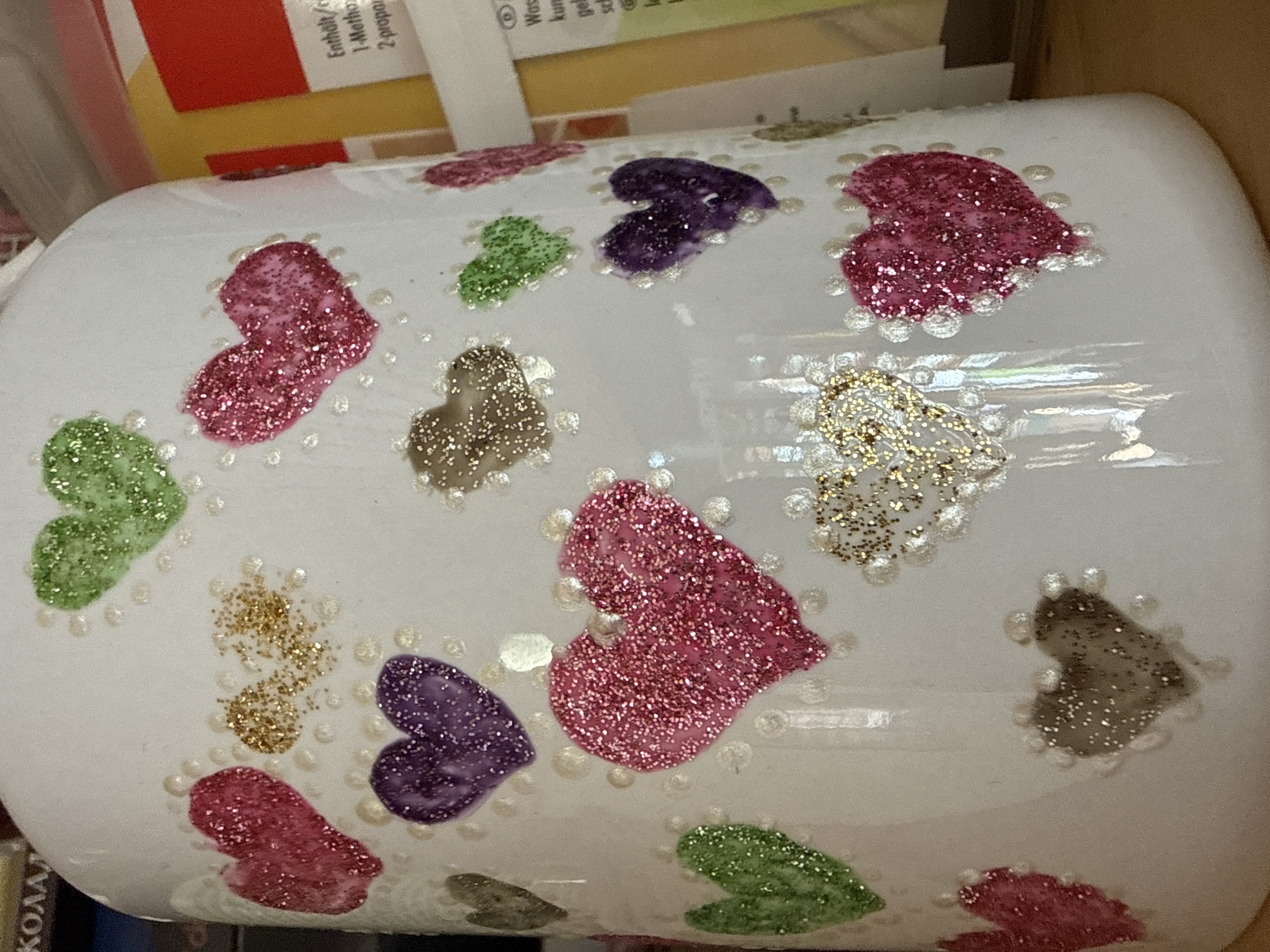 The image depicts a white, cylindrical object adorned with glittery hearts of various colors (pink, purple, green, gold, and gray). Each heart is outlined with small, white, pearl-like beads. The object is surrounded by other items; a red and yellow box is partially visible in the upper left, and part of a book or box with Cyrillic text ("КОЛА") is visible in the lower left corner. The scene suggests a craft project or decorated item. There is no verifiable information to determine if this object is from Troyan, Bulgaria. The location cannot be determined from the visual information provided.