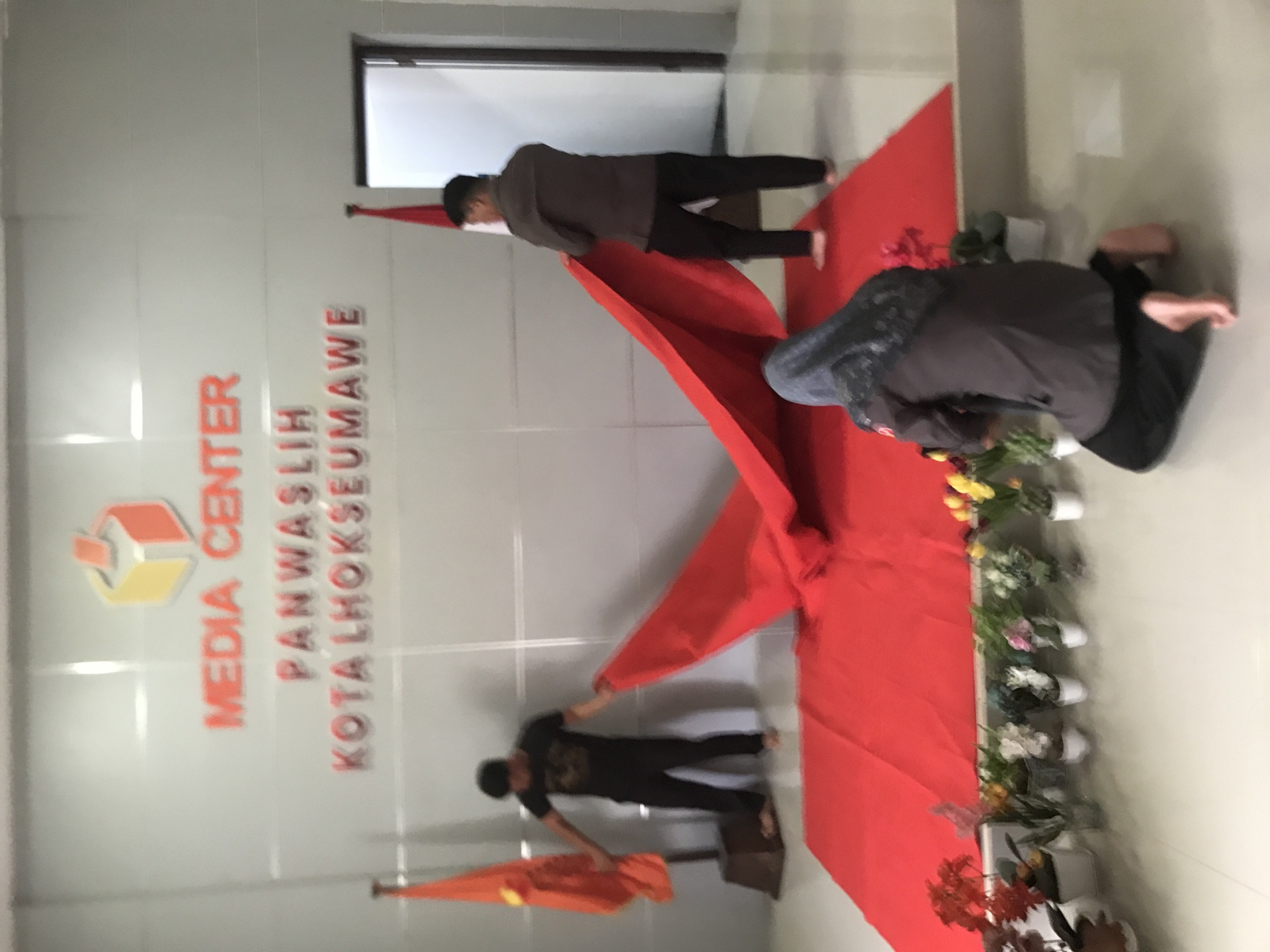 The image shows three people setting up a red carpet for an event at the "Media Center Panwaslih Kota Lhokseumawe" in Lhokseumawe, Indonesia. The environment is a brightly lit interior space with a white wall bearing the organization's name and logo in red. The red carpet is flanked by potted plants with colorful flowers. One person is kneeling on the carpet arranging flowers. The other two people are standing on the carpet in order to set it up correctly.