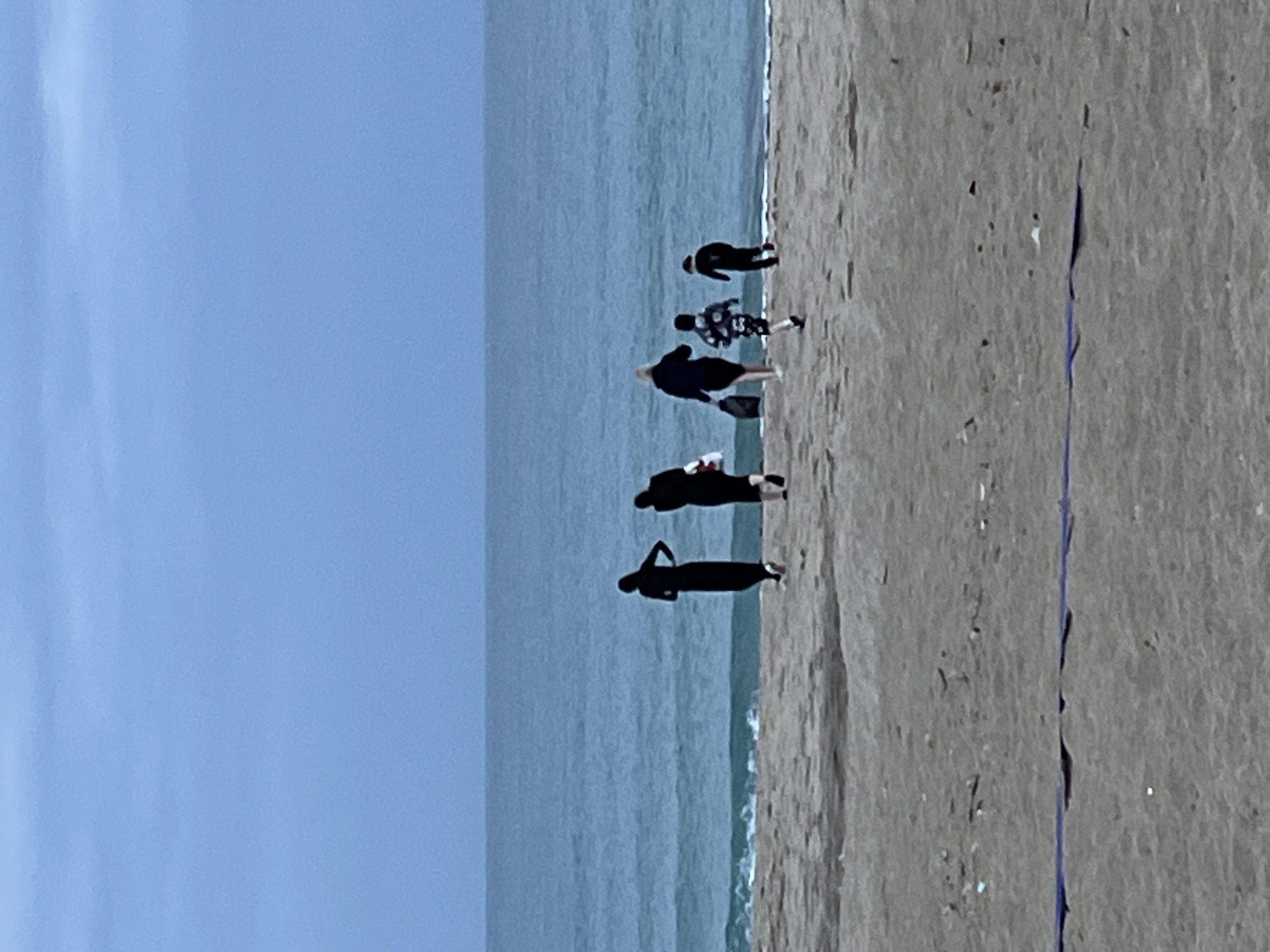 The image shows a beach scene in Alanya, Turkey. The beach is tan, and the water is a muted blue, leading to a light blue sky. Six people are on the beach, seemingly walking along the shoreline. They are grouped, with the person closest to the camera in the bottom of the frame. The image is oriented with the horizon appearing vertically.