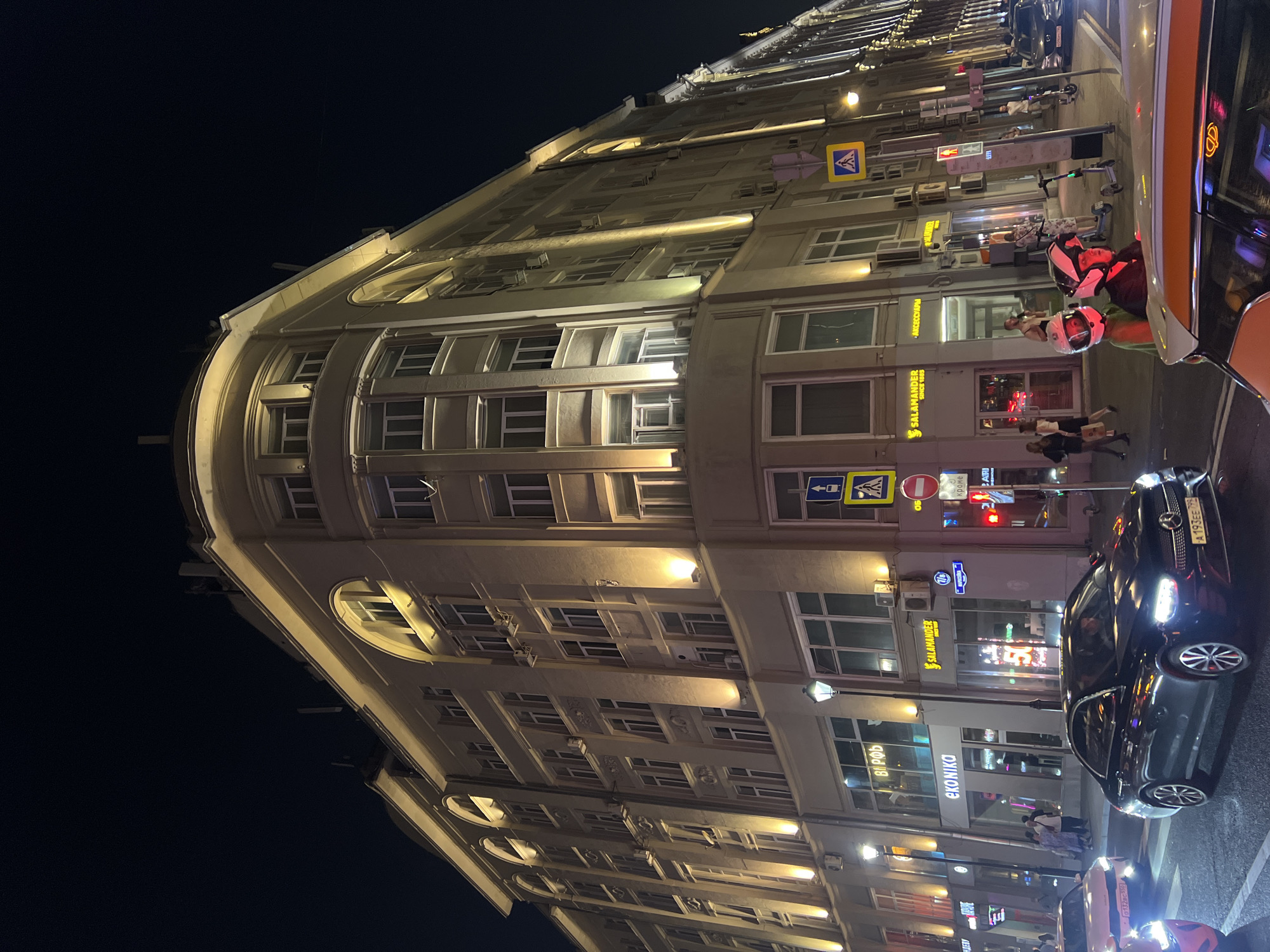 At night, a wide, low-angle shot captures the facade of a multi-story building on a city street in Moscow, Russia. The building, a cream-colored structure with a rounded corner facing the camera, is illuminated by warm, yellow lights that highlight its architectural features, including arched window frames on the upper floors and rows of rectangular windows. A cornice runs along the top of the building, also lit from below. The street below is busy with traffic, including several cars, and pedestrians are visible on the sidewalk. Signs for businesses such as "EKONIKA" and "SALAMANDER" are displayed on the ground floor. Traffic lights and street signs are present. Two people wearing helmets, possibly on a scooter or motorcycle, are in the foreground. The sky above is dark.