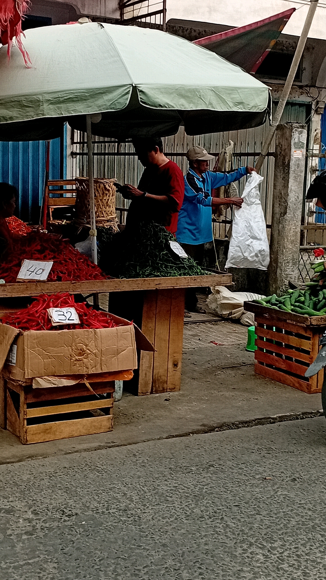 The image depicts a street market scene, likely in Jambi, Indonesia, under a large, light-colored umbrella providing shade. Two men are visible. One man in a red shirt is looking down at a mobile phone. The other man, wearing a blue jacket and a cap, is holding a white plastic bag and interacting with someone to his right (out of frame).
In the foreground, there are piles of red chilies displayed on a wooden table, with price tags indicating "40" and "32". A cardboard box filled with red chilies is also present on the lower left. To the right of the table, a wooden crate is filled with cucumbers. The ground is paved with asphalt. The background shows a corrugated metal wall and some fencing. The overall atmosphere suggests an active marketplace environment.