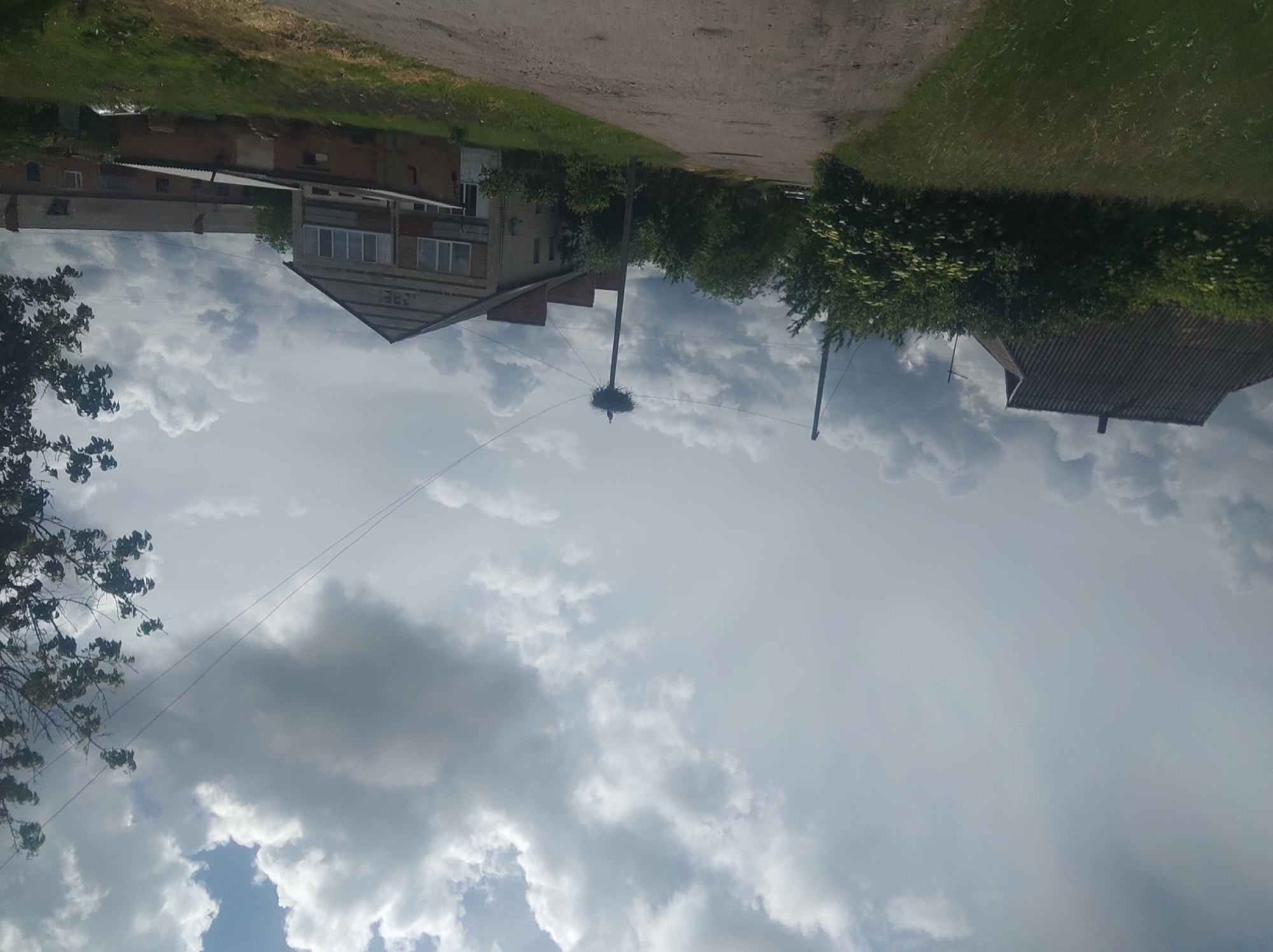A horizontally oriented photograph captures a daytime scene in what appears to be a residential area of Unknown. The sky, filled with billowing gray and white clouds, dominates the view. Below, various buildings line the landscape. On the left, a two-story building with a pointed roof stands next to a longer, lower structure. In the center, a utility pole supports a large bird's nest, possibly a stork's nest, with a bird visible on top. A second utility pole is positioned to the right. The edge of another building is visible on the far right. The foreground features a dirt road and green vegetation, including grass and trees. The photograph is slightly skewed, giving it a tilted appearance.