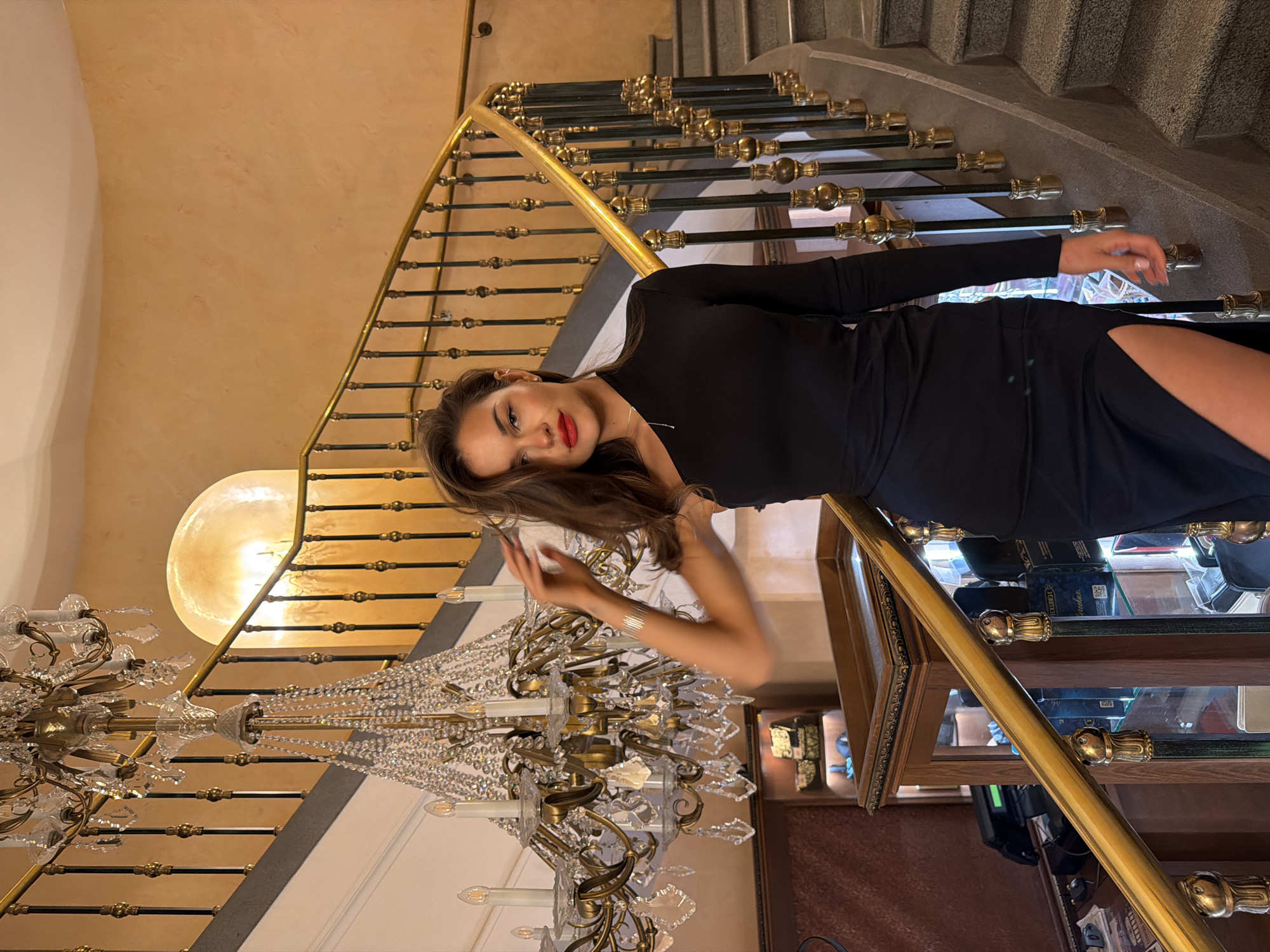 An elegant young woman poses for a photograph on a grand staircase, likely within a luxurious establishment in Moscow, Russia. She has long, dark brown hair and wears a long-sleeved black dress with a high slit, revealing her left leg. Her lips are a vibrant red, and she gazes directly at the camera with a composed expression. She leans against an ornate staircase railing, which features a polished golden handrail and dark balusters adorned with golden decorative elements. The staircase steps appear to be made of light-colored stone or concrete.
The interior showcases a sophisticated and grand design. To the left and behind the woman, a large, multi-tiered crystal chandelier with numerous light sources hangs prominently. Further back, a large, glowing spherical light fixture casts a warm illumination onto the light beige-colored walls. To the right of the frame, a wooden display case with glass shelves is visible, containing various items, including some blue packaging where text is faintly discernible but illegible. The artificial, warm lighting throughout the scene suggests an indoor setting, possibly an evening event or a lavishly lit lobby. The overall ambiance is one of opulence and grandeur.