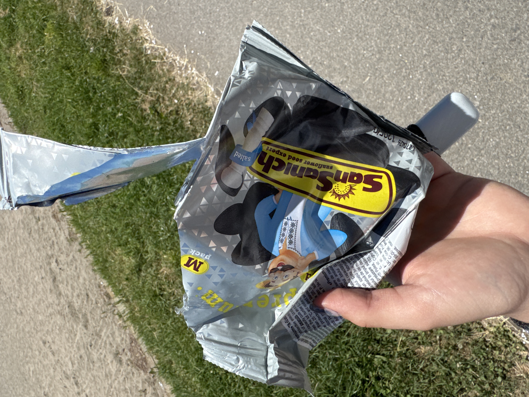 A hand holds a torn bag of "SanSanych sunflower seed expert" snacks. The bag is predominantly silver and black with yellow and blue accents. A cartoon character is depicted on the packaging. The background consists of a patch of green grass adjacent to a paved area, likely a sidewalk or pathway. The scene is set outdoors in the city of Burgas, Bulgaria, on a sunny day.