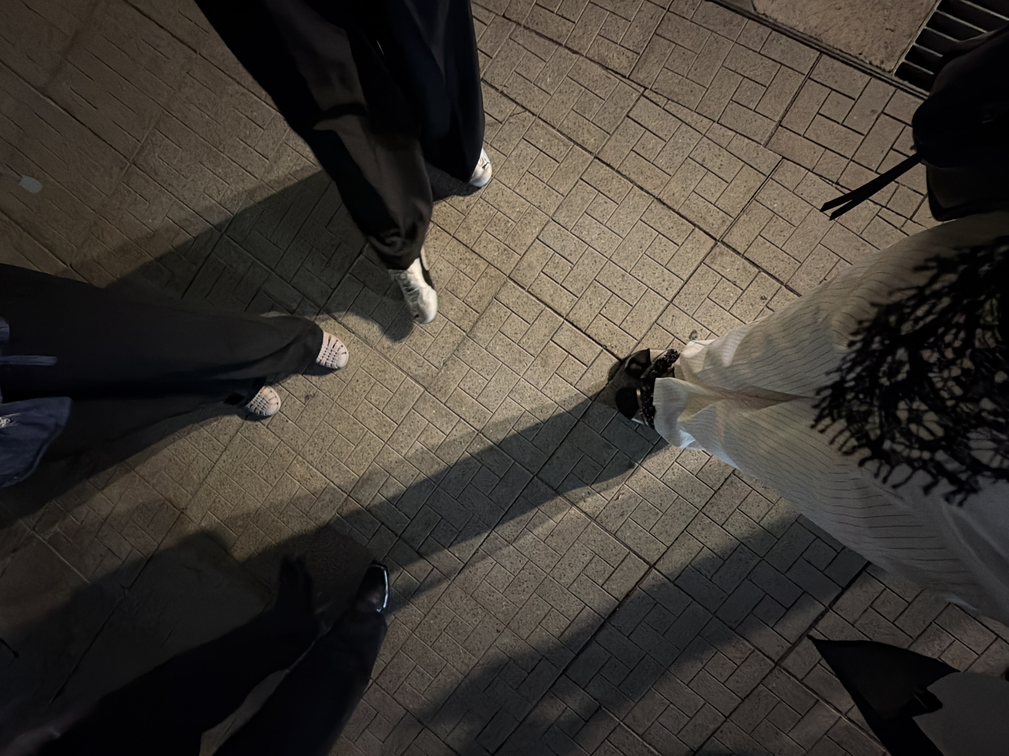 The image shows the lower bodies and legs of at least four individuals standing on a brick-paved sidewalk at night. The lighting suggests artificial light sources, casting long shadows on the ground. The individuals are wearing casual clothing, including dark pants and sneakers. One person is wearing white pants with black pinstripes and a black lace-like design on the front. Another person is wearing white shoes with a woven texture. There is a black umbrella or bag on the left side of the frame and what appears to be a pair of chopsticks or similar thin objects held by one person on the right. The setting is an outdoor urban environment, possibly a public walkway or plaza. The specific location in Bangkok, Thailand cannot be determined from the visual information provided.
