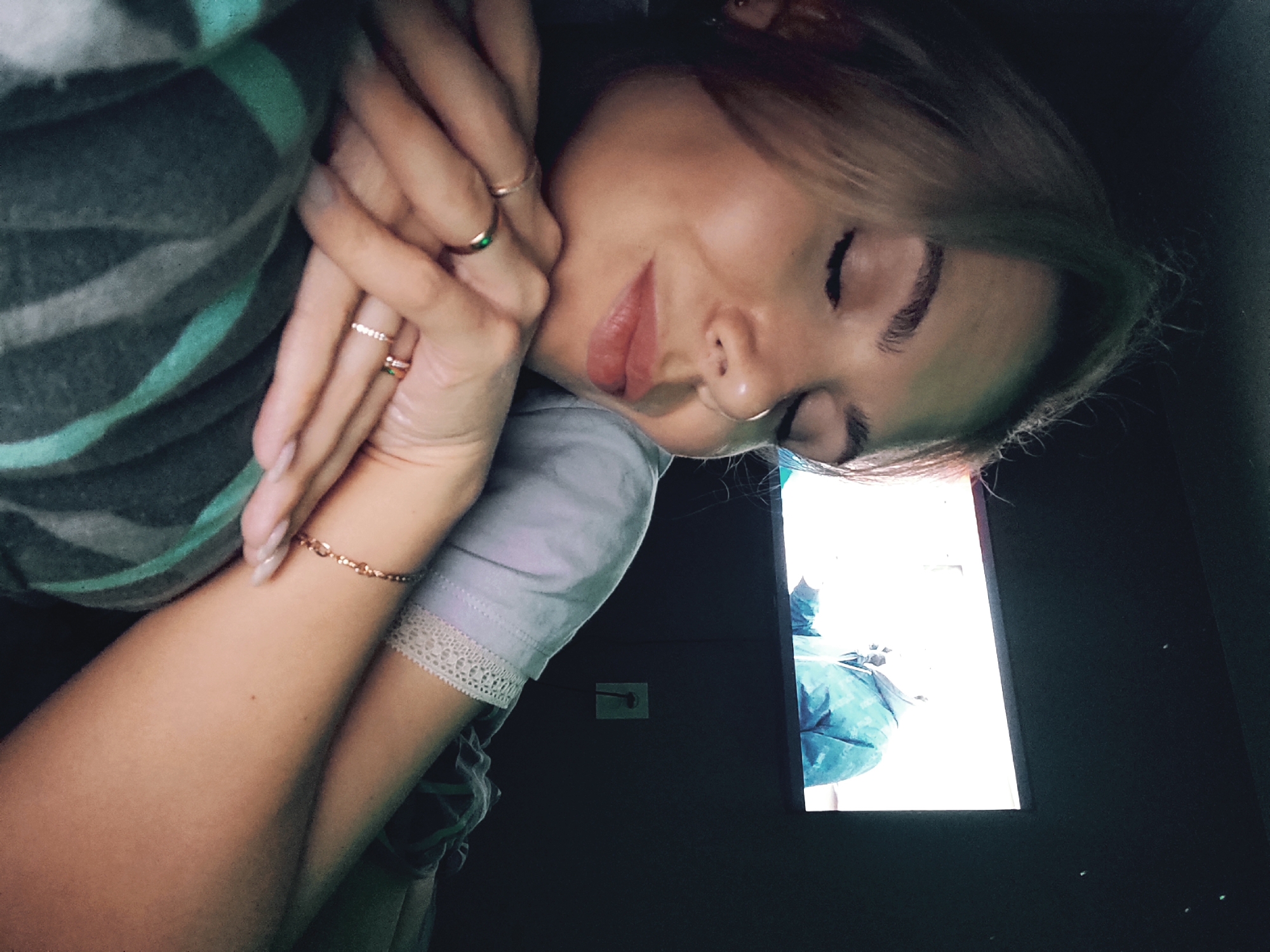 The image presents a close-up, angled portrait of a young woman resting in a dimly lit room in Chaikovsky, Russia, likely at night. She lies on her side with her eyes closed, conveying a sense of peace and relaxation, possibly asleep. Her light brown or blonde hair gently frames her face, and she has full, naturally colored lips, and a small nose piercing.
Her hands are clasped together near her face. She wears multiple rings on her fingers: a thin gold band, another with a small green gemstone, and a sparkling band with tiny stones. A delicate gold chain bracelet adorns her left wrist.
She is dressed in a light lavender or pale purple short-sleeved top with a white lace trim along the cuff. A soft, striped garment or blanket, featuring shades of teal/mint green and dark gray, covers part of her body.
The background is dark, with the primary light source coming from a bright, rectangular screen, likely a television or monitor, positioned to her right. The screen displays a bright, indistinct scene that appears to show a person wearing a blue jacket. On the dark wall below the screen, a white electrical outlet is visible. The atmosphere is quiet and intimate. No clear text is decipherable within the image.