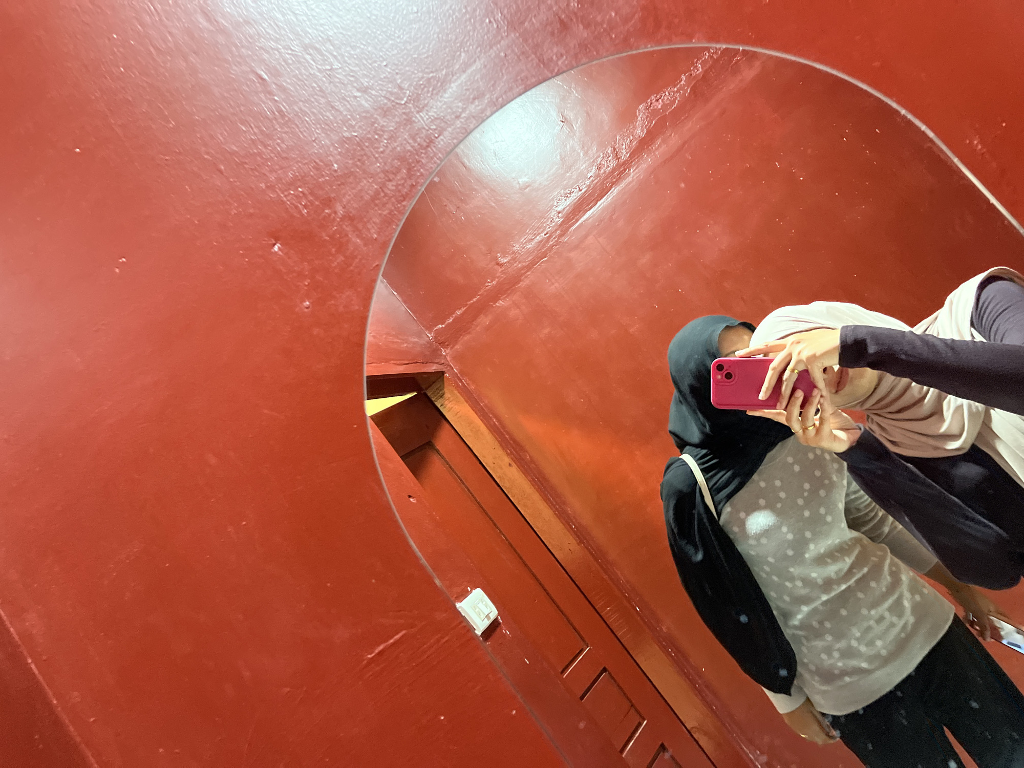A person, wearing a hijab, is taking a mirror selfie in a room with bright red walls. The person is holding a pink smartphone to capture the reflection. They are wearing a light-colored shirt with polka dots, a dark hijab, and dark pants. The reflection shows the person's upper body and face. The background is dominated by the vibrant red color of the walls, which appear to have a slightly textured or worn surface. There are some subtle variations in the lighting, creating highlights and shadows on the wall. The scene is set indoors, and the overall impression is one of a simple, personal moment captured in a visually striking environment. The provided information about the location in Lhokseumawe, Indonesia, is not discernible from the visual content of the image itself.
