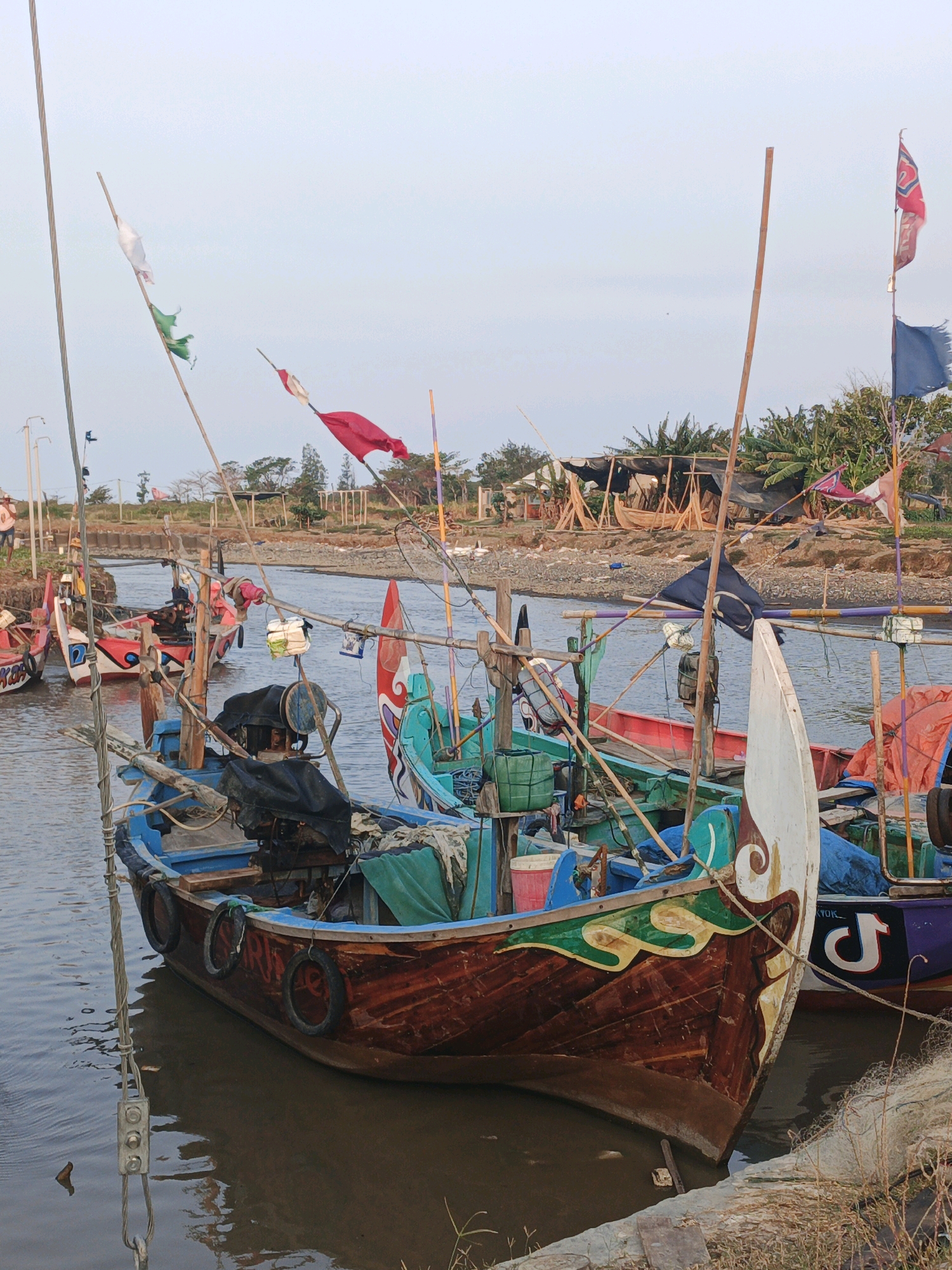 The image depicts a fleet of small fishing boats moored in a narrow waterway in Bumiharjo, Indonesia. The boats are wooden, with brightly colored, intricate paintwork on their prows. Many are equipped with outboard motors and fishing gear, including nets and poles. Several flags, some tattered, are affixed to poles on the boats and along the shore, adding a festive and weathered touch to the scene. The waterway is calm and murky, reflecting the overcast sky. In the background, palm trees and other vegetation suggest a tropical environment. A few wooden structures and what appear to be makeshift shelters are visible on the riverbank, indicating ongoing activity or construction. The scene is one of a working fishing village, bustling with life and tradition.