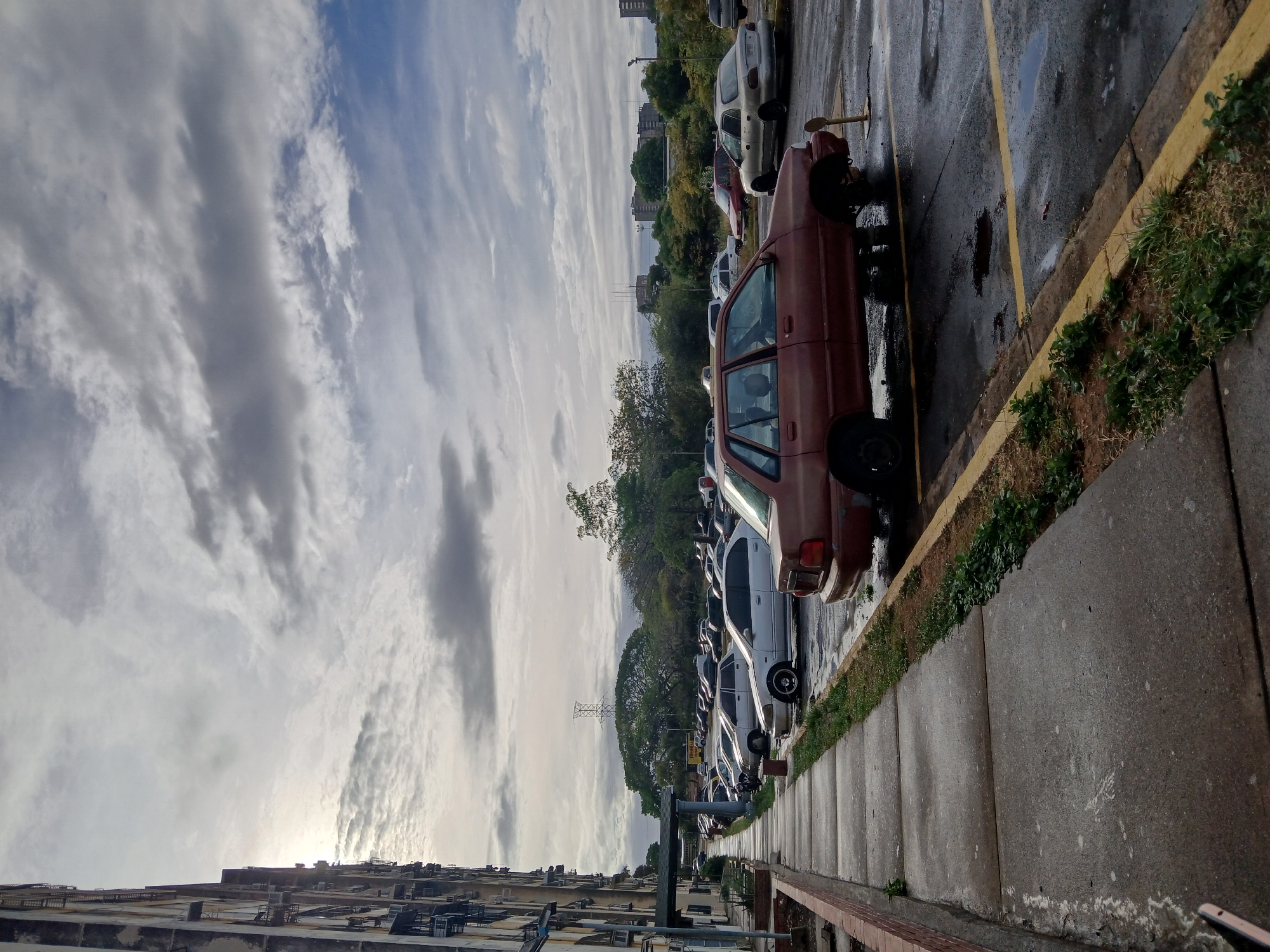 The image captures an outdoor scene in Ciudad Guayana, Venezuela, featuring a multi-story residential building, a parking lot, and an overcast sky. The sun is partially visible behind the building on the left.
A concrete sidewalk runs along the left, bordered by a low red brick wall and a curb with overgrown grass. The asphalt parking lot, displaying wet patches and faded yellow parking lines, occupies the center and right.
In the immediate foreground, a reddish-brown, four-door sedan (consistent with models like a Kia Sephia) is parked. Its front left wheel is missing, and the vehicle is supported by a makeshift wooden block or jack stand. The rear passenger door is open, and the vehicle shows significant signs of weathering and disrepair.
Numerous other sedans, mostly light-colored, are parked in rows throughout the lot. A silver Honda Civic (mid-1990s model) is visible further back, also missing a front left hubcap. No individuals are present in the frame.
Beyond the parking lot, a dense line of trees partially obscures distant urban buildings and a tall, lattice-structure power transmission tower. The multi-story building on the left exhibits an aged concrete facade with visible windows and external AC units or piping. The overall environment presents as an urban residential area with weathered infrastructure.