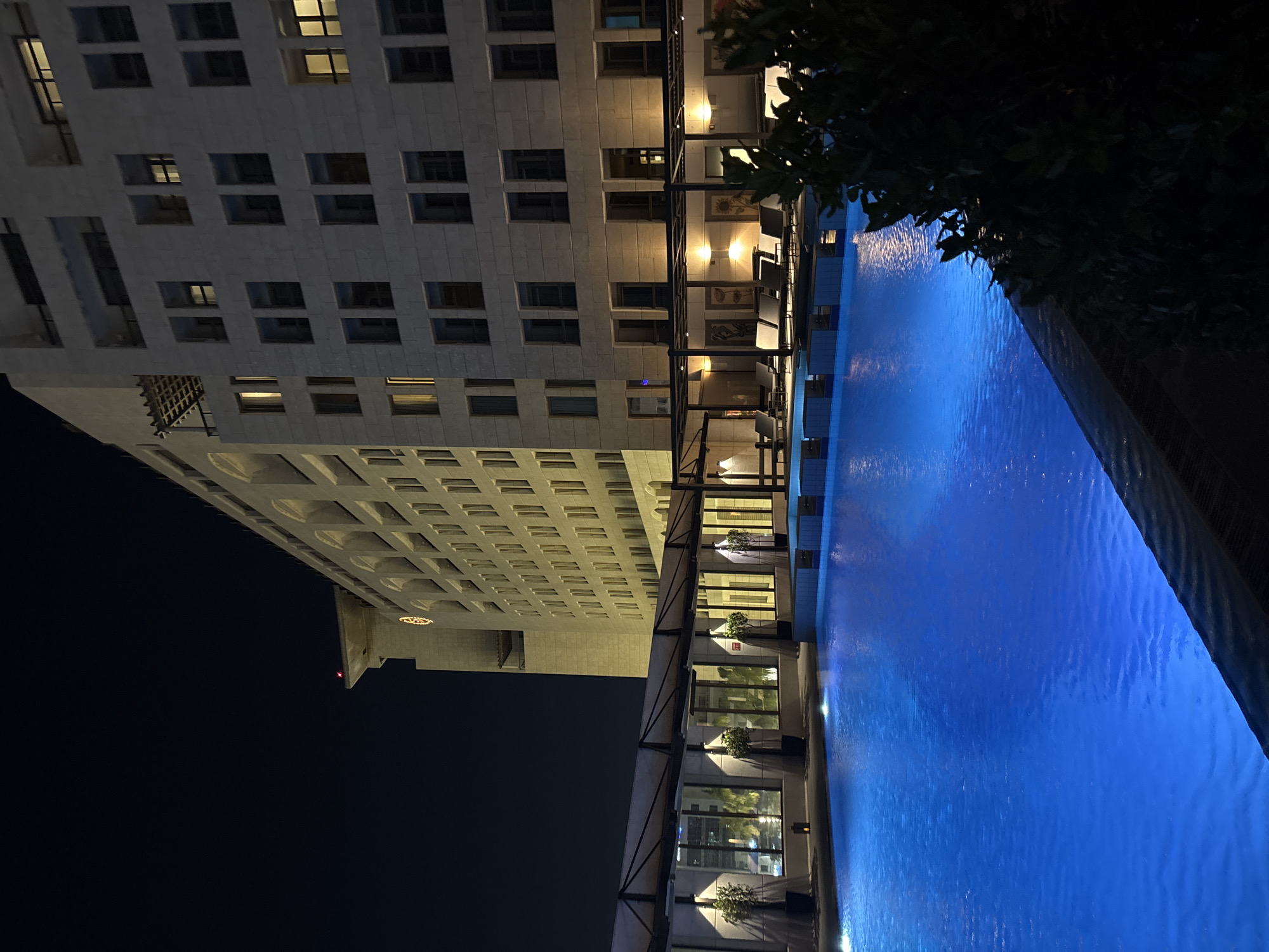 A nighttime scene features a brightly lit blue swimming pool in the foreground. In the background, a large, modern building with many windows, some illuminated, stands tall. The building has a light-colored stone facade and appears to be a hotel or residential complex. There is a covered area with lounge chairs and plants visible along the side of the pool. The sky above is dark and clear. The building's architecture suggests it is located in a developed urban area. Given the architectural style and the mention of Amman, Jordan, this likely depicts a hotel or resort in the city, possibly a luxury establishment with an outdoor pool.