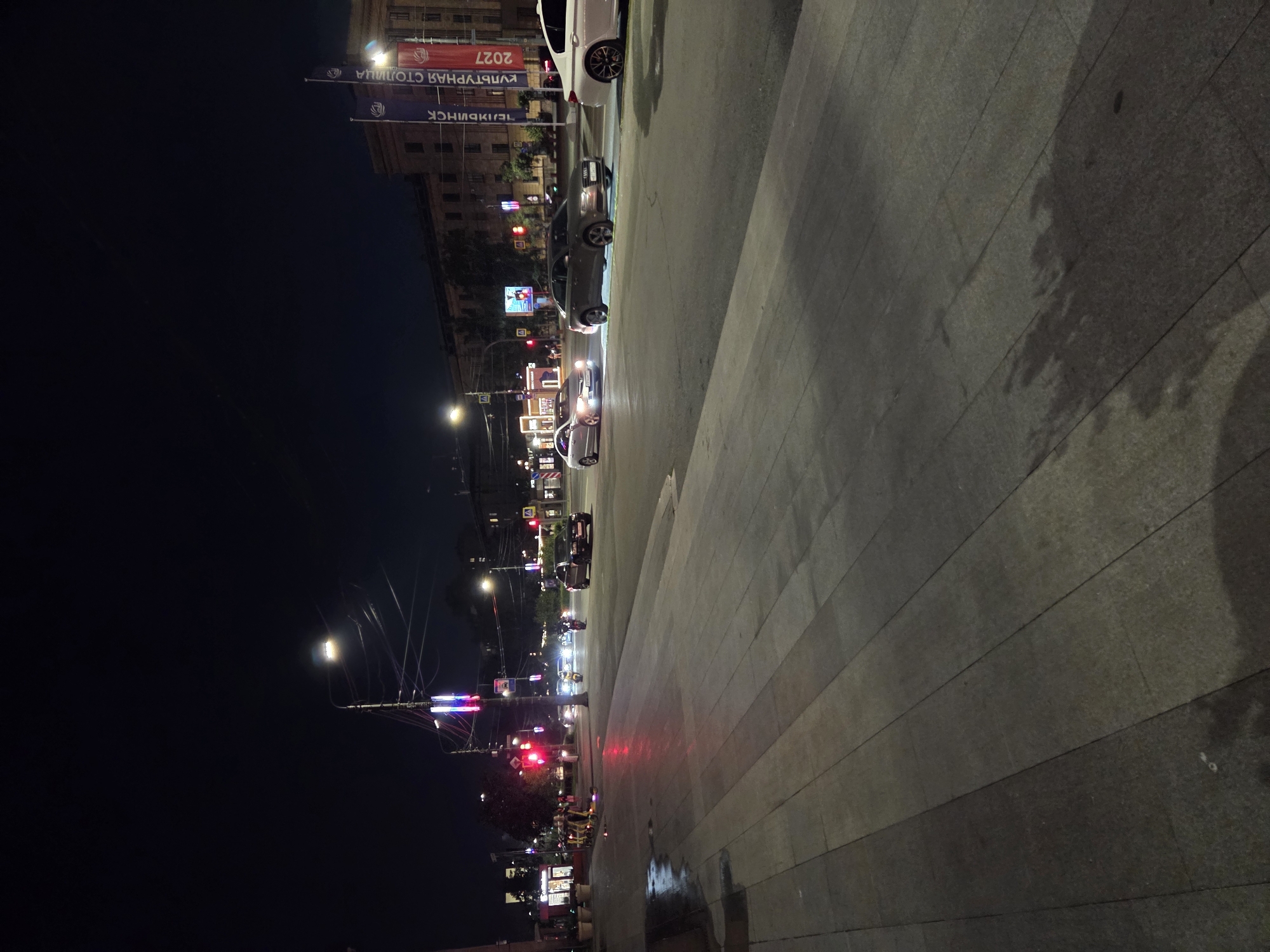 The image depicts a nighttime street scene in Chelyabinsk, Russia. The perspective is from a sidewalk, looking across a wide, multi-lane street. Several cars are visible on the road, some in motion and some stopped at what appears to be an intersection with traffic lights.
In the foreground, the sidewalk is paved with large, grey, rectangular stone slabs, some of which appear wet or reflective, suggesting recent rain or cleaning. There are shadows cast on the sidewalk, likely from trees or buildings not fully visible.
Further into the scene, streetlights illuminate the road and the surrounding buildings. One prominent streetlight with multiple bulbs is visible. There are also brightly lit signs and building facades. Two tall, vertical banners are attached to a building on the right side of the street, one blue and one red, with Cyrillic lettering that appears to read "КУЛЬТУРНАЯ СТОЛИЦА 2027" (Cultural Capital 2027).
Several vehicles are present: a white sedan on the far right, a grey Audi sedan in the middle lane, a silver sedan in the left lane, and various other cars and what appear to be scooters or motorcycles further down the street. One of the scooters in the foreground on the left is yellow.
The overall atmosphere is that of an urban environment at night, with artificial lighting creating reflections and casting shadows. The buildings lining the street appear to be of moderate height and architectural style common to Russian cities.
