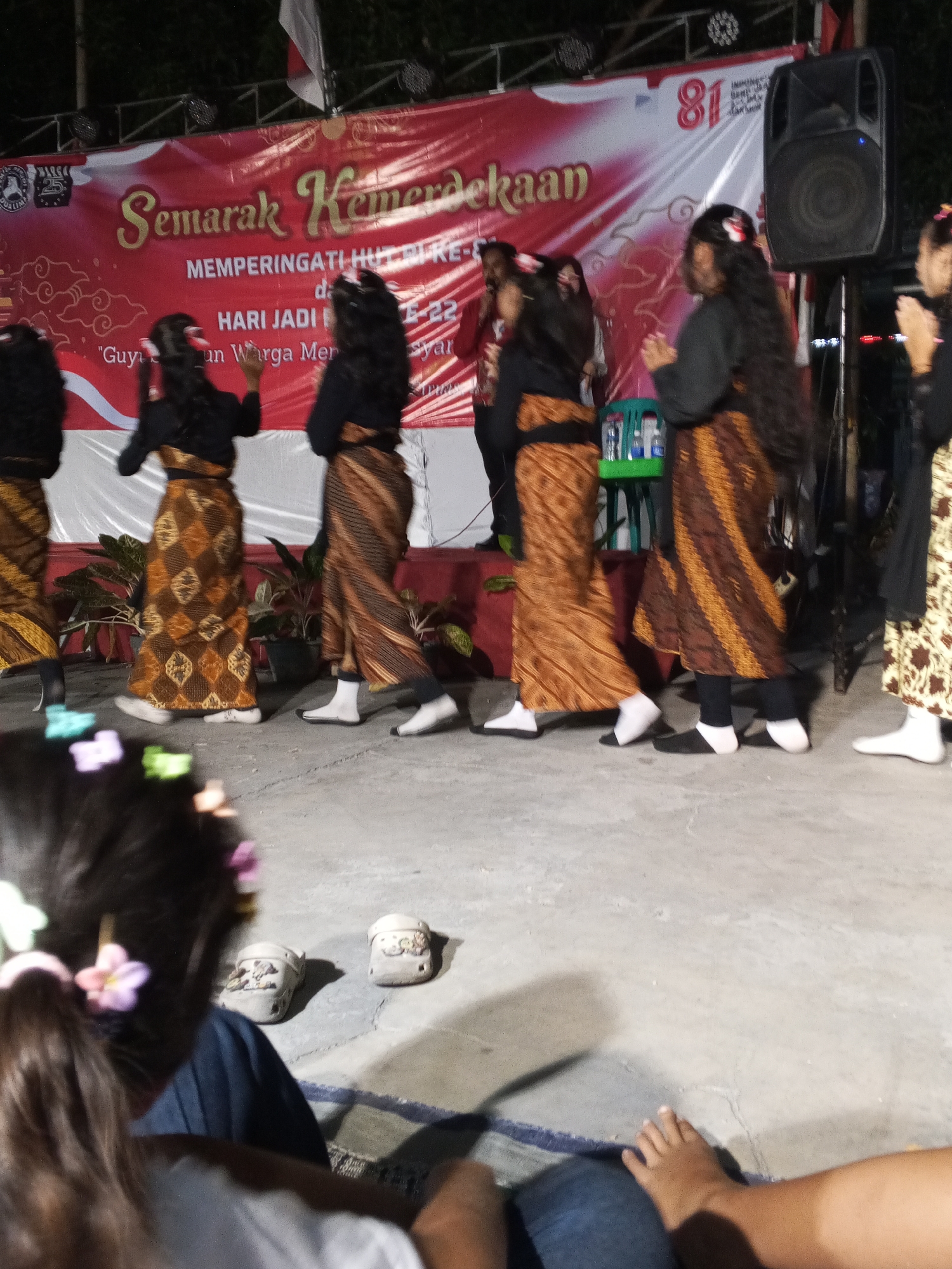 The image depicts an outdoor event at night in what is identified as the city of Ciruas, Indonesia, likely celebrating Indonesian Independence Day, as indicated by the banner reading "Semarak Kemerdekaan" (Independence Festivity) and "Memperingati HUT RI-Ke-77" (Commemorating the 77th Indonesian Independence Day). A group of young women, appearing to be performers, are dressed in traditional Indonesian attire, specifically batik skirts and black tops, with some adorned with floral hair accessories. They are dancing on a concrete ground in front of a stage draped with a large red and white banner. The stage is illuminated by lights, and a speaker is visible to the right. In the foreground, a child's back is visible, facing away from the camera, with colorful hair clips. Two pairs of children's shoes, possibly Crocs with cartoon characters, are placed on the ground in the lower left quadrant. The overall scene suggests a community celebration.