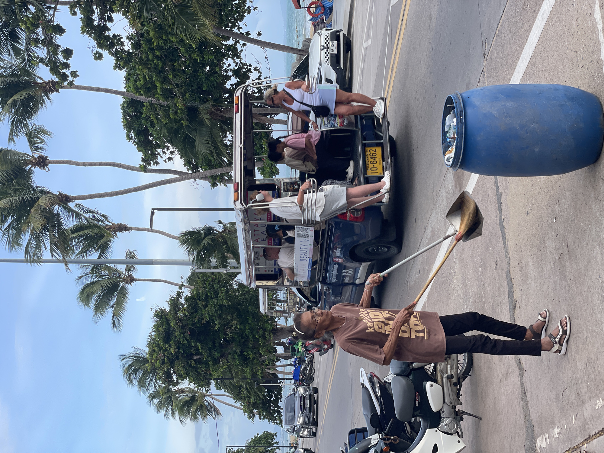 In Pattaya City, Thailand, an elderly woman with glasses and a brown t-shirt is sweeping debris with a shovel into a blue barrel on the sidewalk. She is wearing black pants and sandals. In the background, a blue pickup truck modified with a covered seating area for passengers is parked on the road. Several people are seated in the truck. A white car is parked on the right side of the road, and several motorcycles are parked on the left side. Palm trees and lush green trees line the street, with the ocean visible in the distance. The sky is cloudy and pale blue.