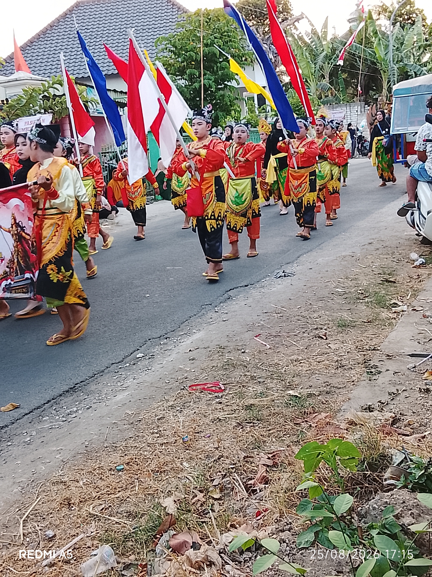 In Semanding, Indonesia, on August 25, 2023, at 17:15, a parade is underway on a paved street. A group of young people, dressed in traditional attire consisting of orange tops, patterned skirts, and yellow waist sashes, are marching and carrying flags. The flags are a mix of red, white, and blue, and some are adorned with a red and white motif. Behind the flag bearers, other participants in similar colorful costumes are visible, along with onlookers. The street is lined with trees and buildings, suggesting a residential or commercial area. The ground beside the street is covered with dry grass and scattered debris, indicating a somewhat unkempt environment. A white vehicle, possibly a motorcycle, is parked on the right side of the frame, and a person is seated on it, observing the parade. The overall scene depicts a cultural event or celebration in a rural or semi-urban setting.