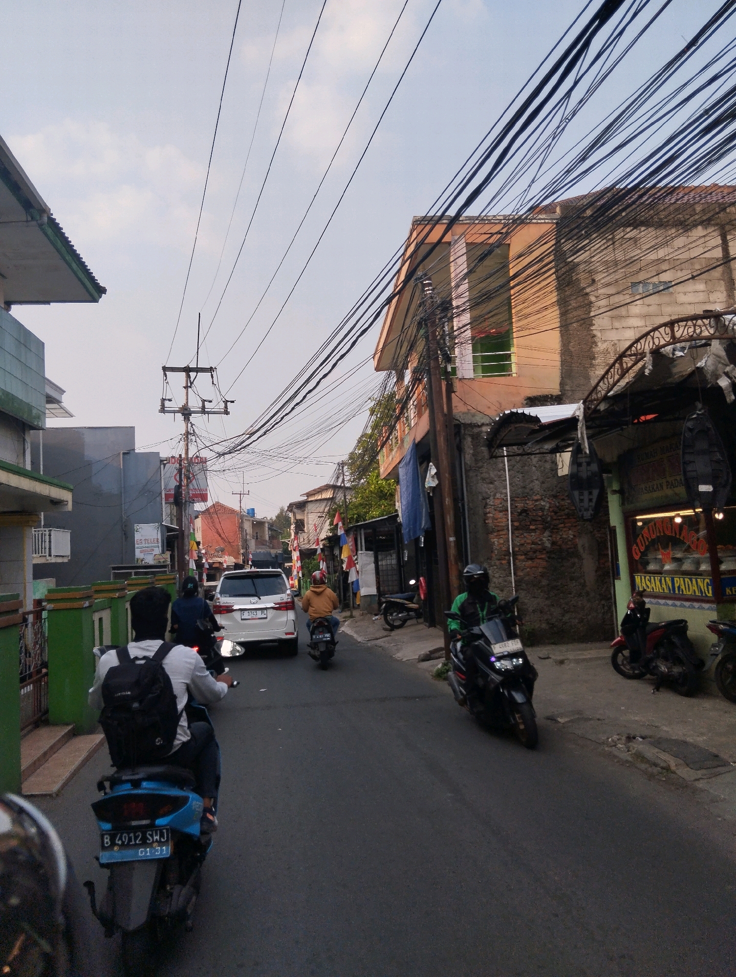 The image depicts a street scene in Sukaraja, Indonesia, with several motorcycles and a white car navigating the road. The street is lined with buildings, some appearing to be residential and others commercial. Overhead, a dense network of electrical wires crisscrosses the sky, creating a cluttered yet characteristic urban aesthetic.
In the foreground, a person on a blue scooter wearing a white shirt and a black backpack is riding down the street. Behind them, a white car with license plate "F 1743 PL" is visible. Further down the road, another scooter rider in a yellow jacket is approaching, and to the right, a person on a black scooter wearing a green jacket is moving away from the camera. Several other motorcycles are parked or in motion along the street and sidewalk.
The buildings display a mix of architectural styles, with some showing signs of age and others appearing more modern. One building on the right has a sign that reads "GUNUNG AGUNG MASAKAN PADANG," indicating a restaurant. The sky is overcast with some visible clouds. The overall impression is of a busy, everyday street in an Indonesian city.