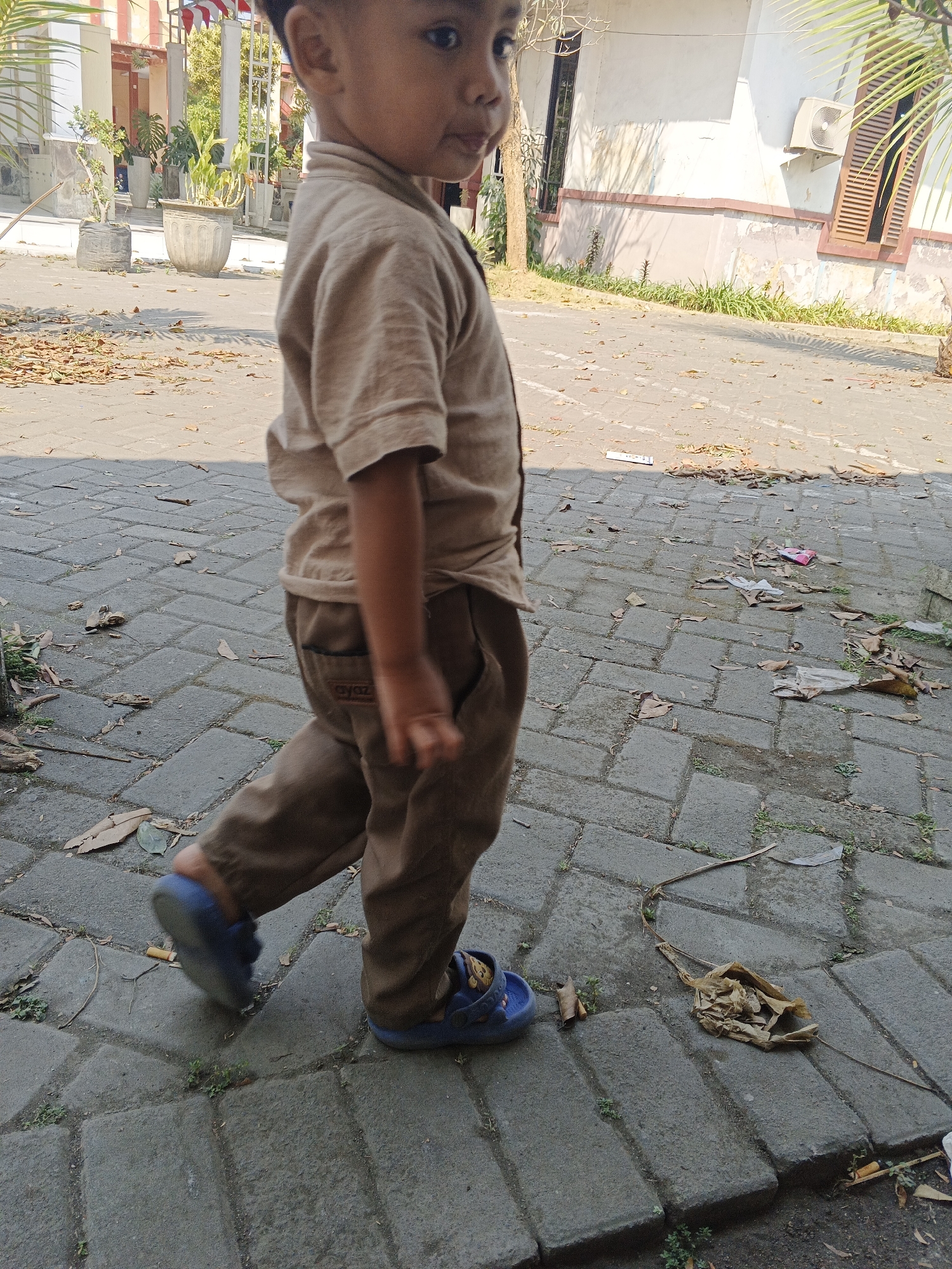 A young boy, wearing a tan short-sleeved shirt and tan pants, with blue sandals, walks on a paved pathway. The pathway is made of interlocking grey rectangular bricks. Scattered on the pathway are fallen dry leaves and some pieces of trash, including plastic wrappers and crumpled paper. In the background, there is a building with white and pink walls, windows with brown wooden shutters, and an air conditioning unit mounted on the wall. Several potted plants are visible in front of the building. The scene appears to be an outdoor area, possibly a street or a schoolyard, in Krian, Indonesia, suggested by the presence of tropical foliage and the architectural style of the building. The boy is looking to his right as he walks, with his left leg forward. The lighting suggests it is daytime, with sunlight casting shadows.