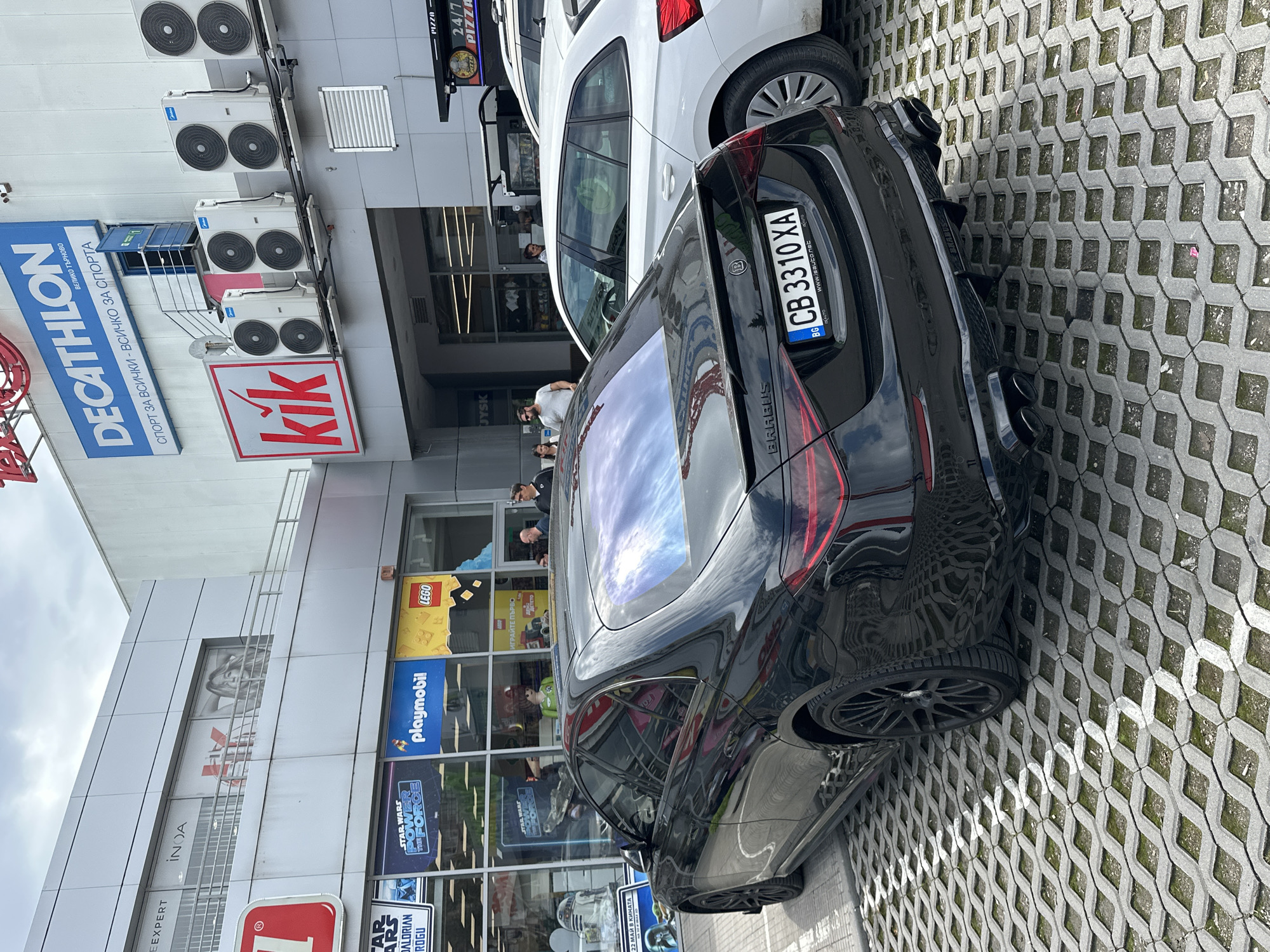 The image shows a black Brabus car parked in front of a shopping center in Veliko Tarnovo, Bulgaria. The license plate of the car is CB 3310 XA. The shopping center features stores such as Decathlon, Kik, JYSK, a Pizza 24/7 restaurant, and a toy store with LEGO and Playmobil displays. Several people can be seen entering or exiting the shopping center. The parking area is paved with interlocking concrete blocks.