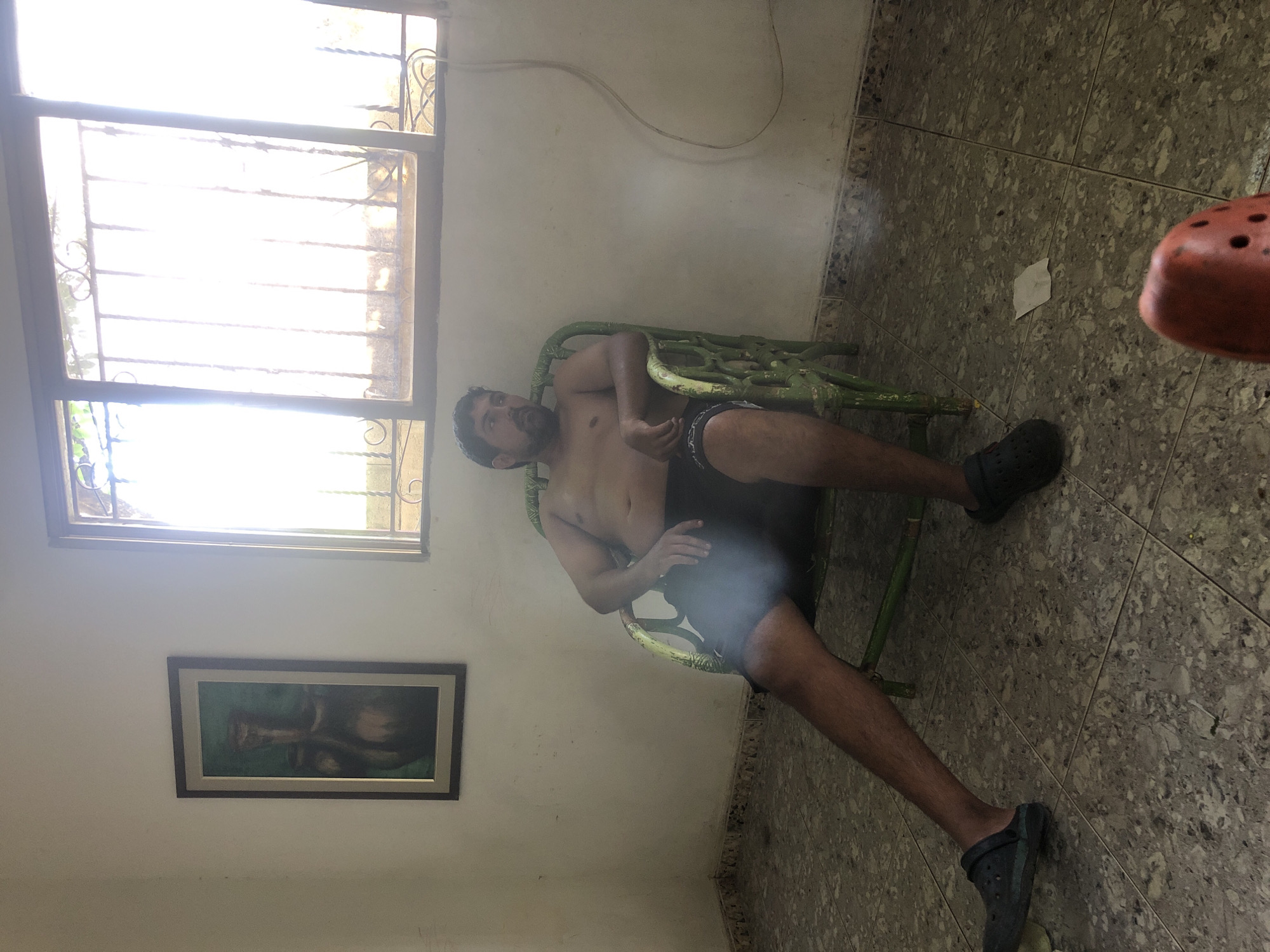 A shirtless man with a beard, wearing black shorts and dark clogs, is seated in a worn, green metal chair in an indoor room. He is looking away from the camera towards a window. The room has white walls, a tiled floor, and a framed painting on the left wall. A large window with bars is visible on the right, letting in bright light. There is a white electrical cord running down the wall. The scene appears to be in a residential setting in La Tagua, Venezuela.