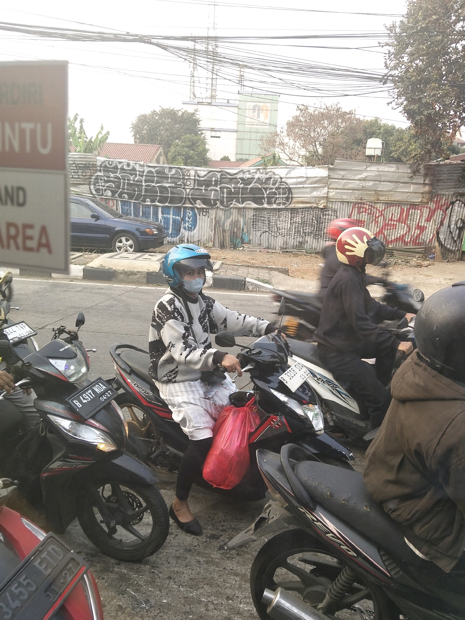 In South Jakarta, Indonesia, on a city street, several motorcycles are stopped. A person wearing a blue helmet and a face mask is seated on a black and white motorcycle, holding a red plastic bag. Another motorcycle is seen with a person wearing a red and yellow helmet. In the background, a blue car is parked, and a corrugated metal fence covered in graffiti lines the street. Overhead, a network of electrical wires is visible against a hazy sky.