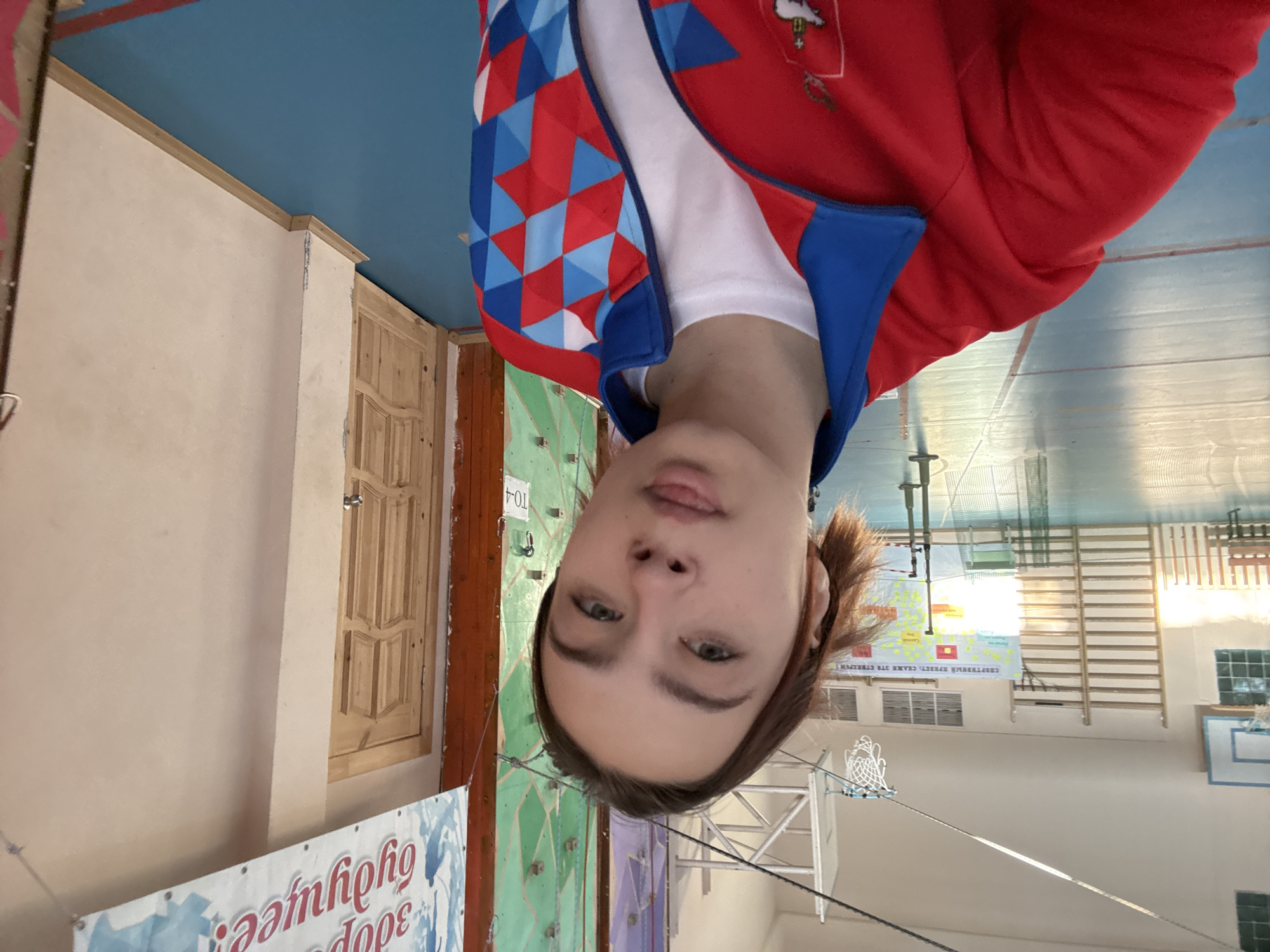 The image shows a person in a red and blue jacket standing inside what appears to be a gymnasium or sports hall. The hall has wooden climbing walls, wall bars, a basketball hoop, and a blue ceiling. A banner with Russian text is partially visible. This photograph might have been taken in the city of Berezniki, Russia.