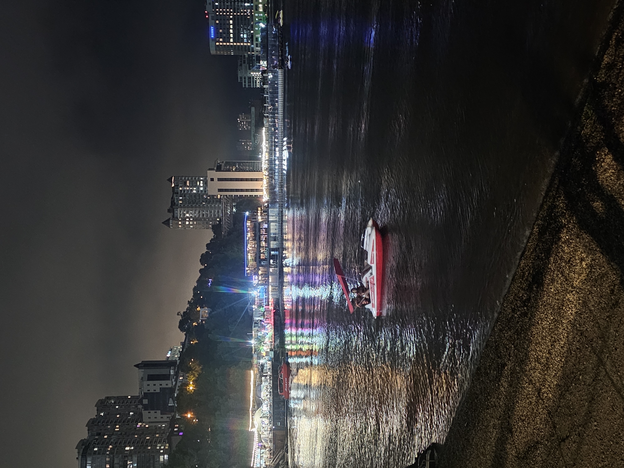 This is a nighttime photograph of a waterfront scene in Vladivostok, Russia. The foreground shows a textured, gravelly embankment leading to dark water. In the middle ground, a small red and white pedal boat with a canopy is on the water, carrying two people. Further out on the water, several other small boats and watercraft are visible, some with lights. The background features a city skyline with illuminated buildings, including high-rise residential structures and commercial buildings with signs. The waterfront itself appears to be a developed area with restaurants or entertainment venues, indicated by numerous colorful lights reflecting on the water. The sky is dark and overcast.