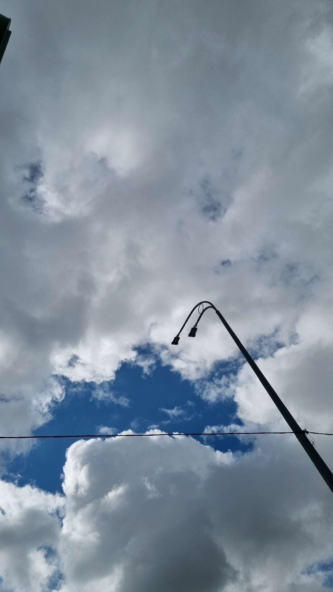 A low-angle shot captures a silhouette of a streetlamp against a dramatic sky filled with a mix of bright blue patches and heavy, gray clouds. The streetlamp features a curved arm with two lights. A thin, dark wire runs horizontally across the lower portion of the image. The scene is set in Dubno, Ukraine.