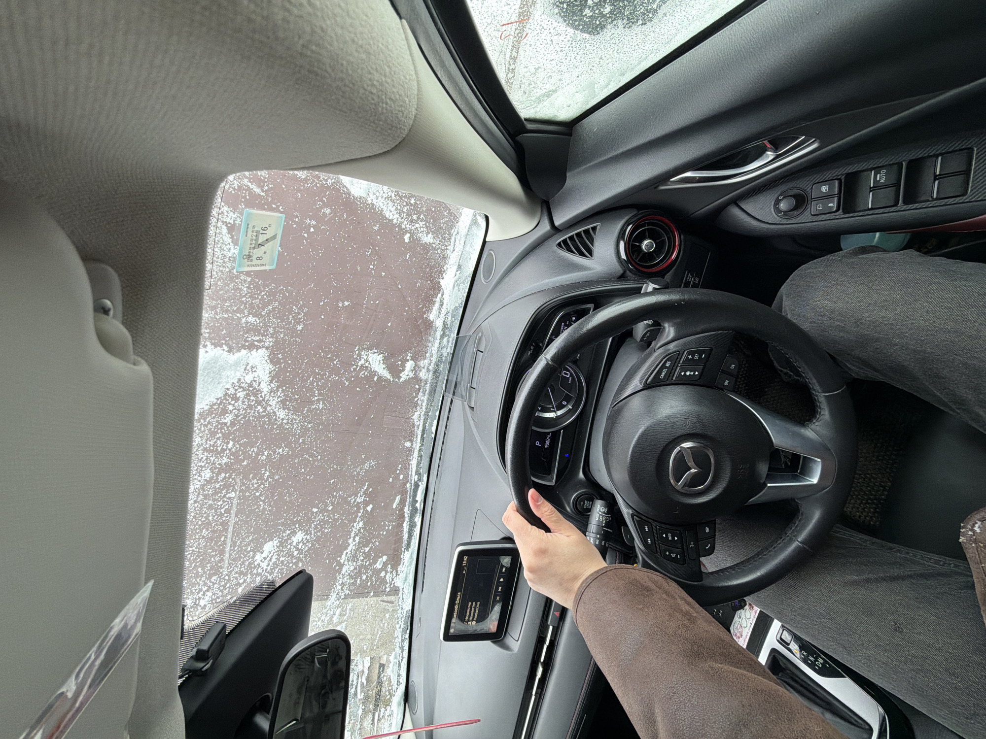 A view from inside a Mazda car, looking out through the windshield which is partially obscured by snow. The driver's hand is visible on the steering wheel. The dashboard includes a built-in display screen and various controls. A parking permit with the number 8 7 16 is affixed to the windshield in the upper left corner. Through the windshield, a paved surface covered with snow is visible. The image, based on the parking permit, is presumed to be in Chaikovsky, Russia. The person is sitting in the driver's seat, holding the steering wheel.