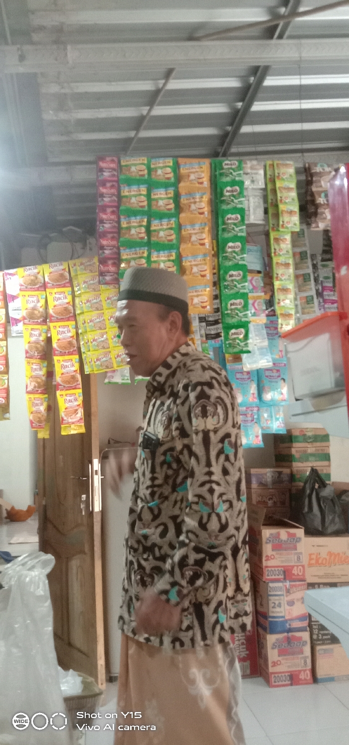 An indoor scene within a small retail establishment or 'warung' in Tarokan, Indonesia. A single adult male is depicted in profile, facing right, positioned centrally. He is an older individual, wearing a light-colored peci cap and a patterned, long-sleeved shirt, possibly batik. His mouth is slightly open. The background is filled with various packaged goods, including sachets of "Energen," "Milo," "Nutrisari," "Masako," and "Racik" products, displayed hanging from overhead lines. To the right, stacks of cardboard boxes containing instant noodles (e.g., "Sedaap," "EkoMie") are visible on the floor. A wooden door with a silver handle is partially visible to the left of the male. A large clear plastic bag containing white contents is on the floor to the far left. The ceiling is constructed of corrugated metal sheets with visible support beams. The floor is light-colored.