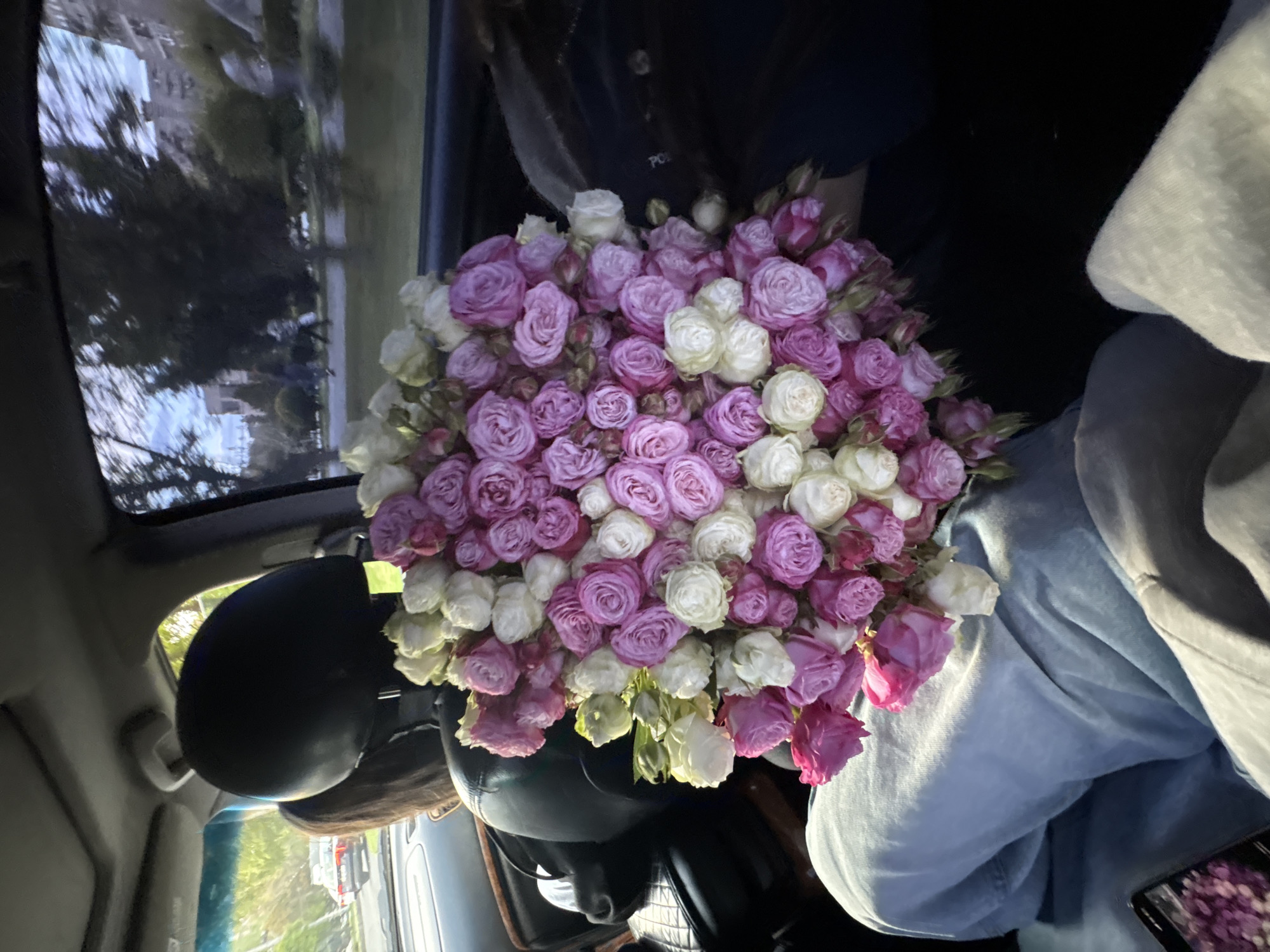 A large, vibrant bouquet of small roses dominates the foreground, held inside a car. The roses are a mix of white/cream and various shades of pink and light purple, densely packed together, suggesting a lush and abundant arrangement.
Two individuals are partially visible within the car. One person, wearing a light blue denim shirt and possibly a light-colored hoodie or jacket, holds the bouquet on their lap or chest. A smartphone is resting on their lap, displaying an image that appears to be more flowers. The second person, likely the driver, is partially obscured behind the bouquet and a black helmet, which rests on the dashboard or seat next to them. Their hair is dark brown.
Through the car windows, particularly the windshield and side window, reflections of a clear daytime sky, green trees, and distant urban or suburban elements are visible. These reflections suggest the car is either moving or parked outdoors on a street in a location like Yerevan, Armenia. Another vehicle, possibly a red or orange truck or car, can be faintly seen in the reflection.
The time of day appears to be daytime, possibly late afternoon, indicated by the soft, ambient light. The weather seems clear and bright. On the dark top worn by the person in the background, a faint and indistinct light-colored text or logo is barely discernible. The overall impression is one of a cheerful occasion, centered around the beautiful floral gift.