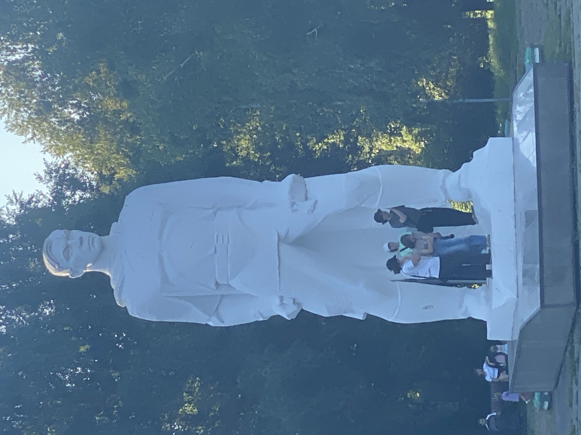 A large, white, monolithic statue of a male figure stands in a park in Novokuznetsk, Russia. The statue is depicted in a military-style uniform and is positioned on a pedestal. Several people are standing at the base of the statue, some appearing to be tourists or visitors. The statue is surrounded by lush green trees, suggesting a warm, sunny day. In the background, other people are visible, as well as what appears to be a playground or recreational area. The overall scene suggests a public gathering or a visit to a memorial site.