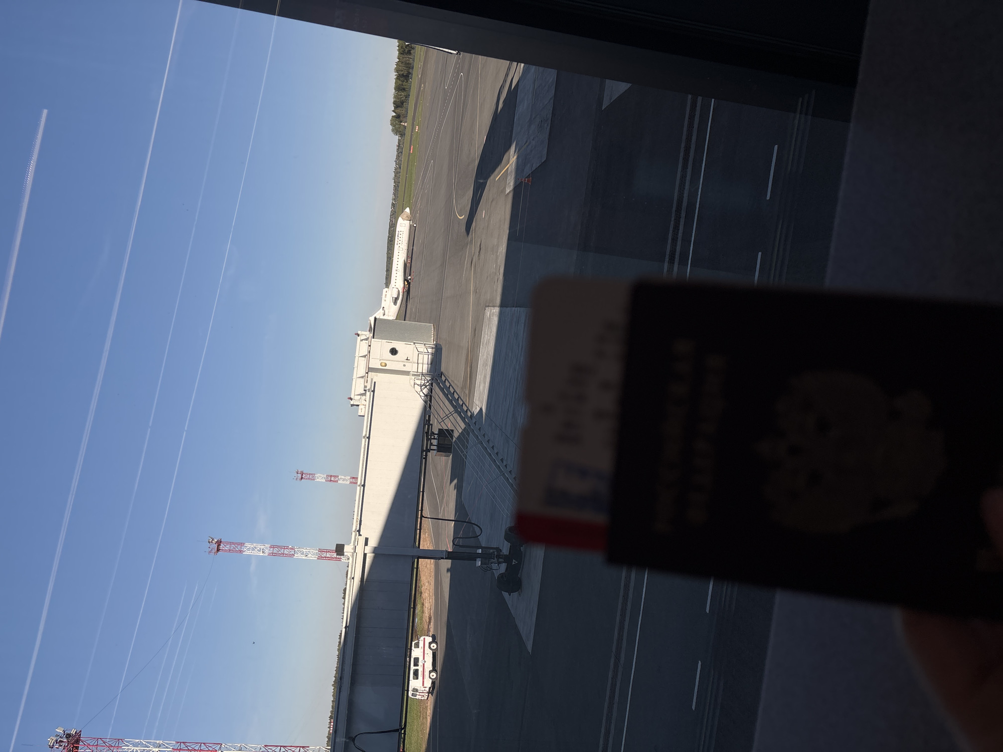 The image depicts an airport scene at Новое Мартьяново, Russia. An airplane is visible on the tarmac, connected to a passenger boarding bridge. In the foreground, a blurred passport is held up against a window. Outside, one can see a clear blue sky marked with contrails, airport cranes, service vehicles, and airport infrastructure. The reflection of the inside of the airport terminal is visible in the window.
