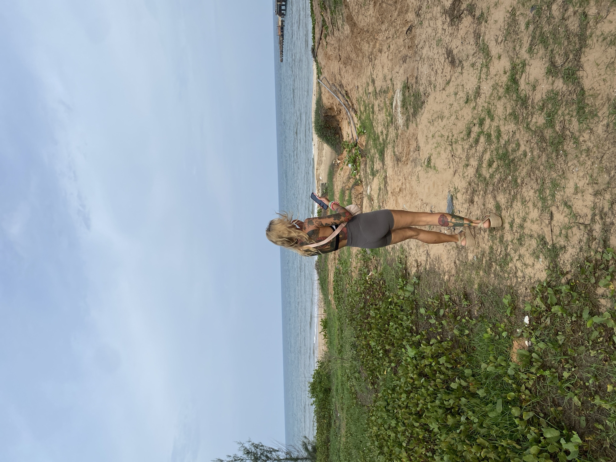 The image depicts a woman standing with her back to the viewer, looking at her smartphone, on an elevated sandy path overlooking a beach and the ocean. She has light-colored hair and is dressed in dark shorts, a patterned top, and light-colored wedge sandals. Visible tattoos adorn her right arm and calf.
The scene is a coastal landscape, identified as Tien Thanh Ward, Vietnam. A calm, light blue ocean stretches across the middle of the frame, meeting a narrow strip of sandy beach below an eroded sandy embankment. A wooden pier or structure is faintly visible extending into the water in the upper right background. The path where the woman stands is composed of dry sand and dirt, interspersed with patches of green grass and leafy vegetation, which becomes denser in the foreground and along the lower edge of the image. The sky is overcast with a soft, light blue hue, suggesting either an overcast day or a time of day with diffused light, such as morning or late afternoon. No visible text is present in the image.