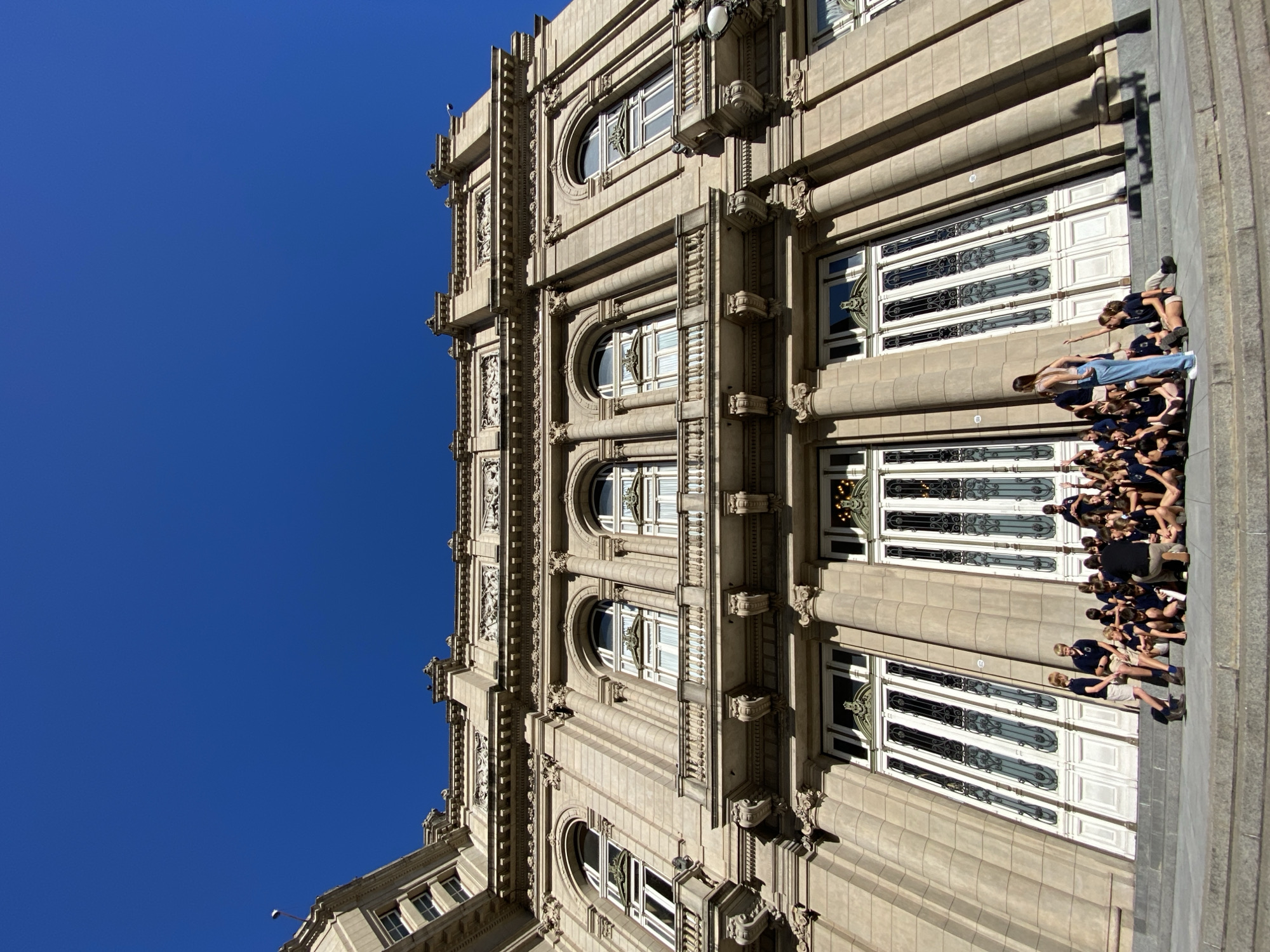 The image displays a grand, multi-story building constructed from light-colored stone, seen from a low-angle perspective against a clear, vibrant blue sky. The building features ornate architectural details, including decorative cornices, pilasters, and intricate carvings, with numerous windows of varying shapes, some arched and others rectangular, often adorned with decorative grilles and balconies. The style appears to be a classical revival or possibly Art Deco, showcasing a sense of grandeur.
At the base of the building, on what appears to be a wide stone plinth or staircase, a large group of people is gathered. Most are young adults or teenagers, predominantly dressed in dark blue or black uniforms, suggesting a school group, tour, or organized event. They are seen standing, sitting, and interacting, with some looking towards the building while others are engaged in conversation. One person in light blue jeans stands out among the uniform-clad crowd.
The scene takes place on a bright, sunny day, indicated by the crisp blue sky and the distinct shadows cast on the building's facade, suggesting mid-morning or afternoon. There are no clouds visible. The overall atmosphere appears calm and organized, typical of a group outing. Given the context of Buenos Aires, Argentina, this is likely a prominent public or historical building. No discernible text is clearly visible on the building or among the individuals.