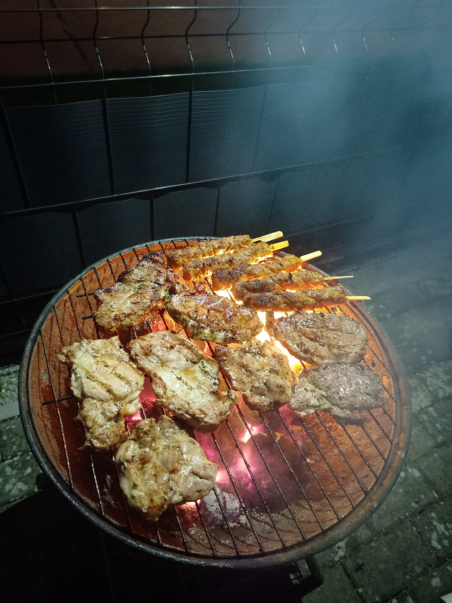 A barbecue grill is depicted outdoors at night in the city of Unknown, Unknown. The grill is round and rusty, filled with hot coals glowing orange and red. On the grill, there are various types of meat cooking, including skewers and what appear to be cuts of chicken and steak. Smoke is visible rising from the grill. A dark fence is in the background. The ground is made of paving stones.